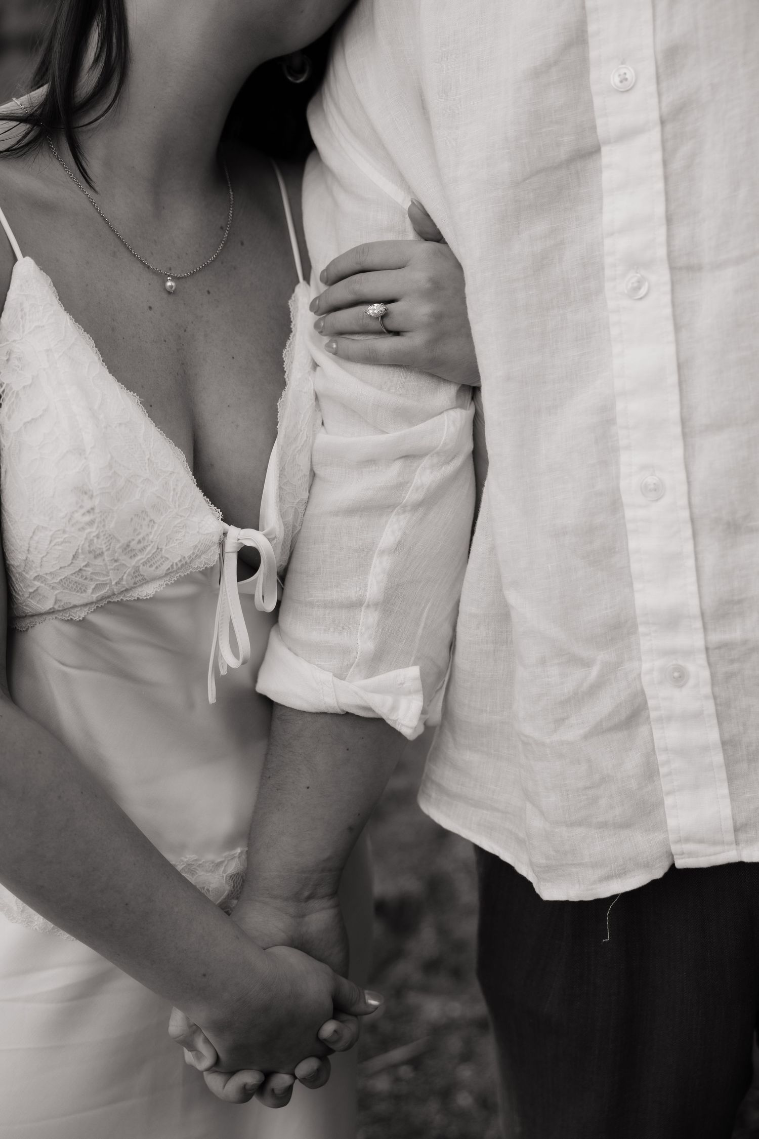 Black and white photo of a couple holding hands, bride wearing lace dress with ring visible, groom in linen shirt.