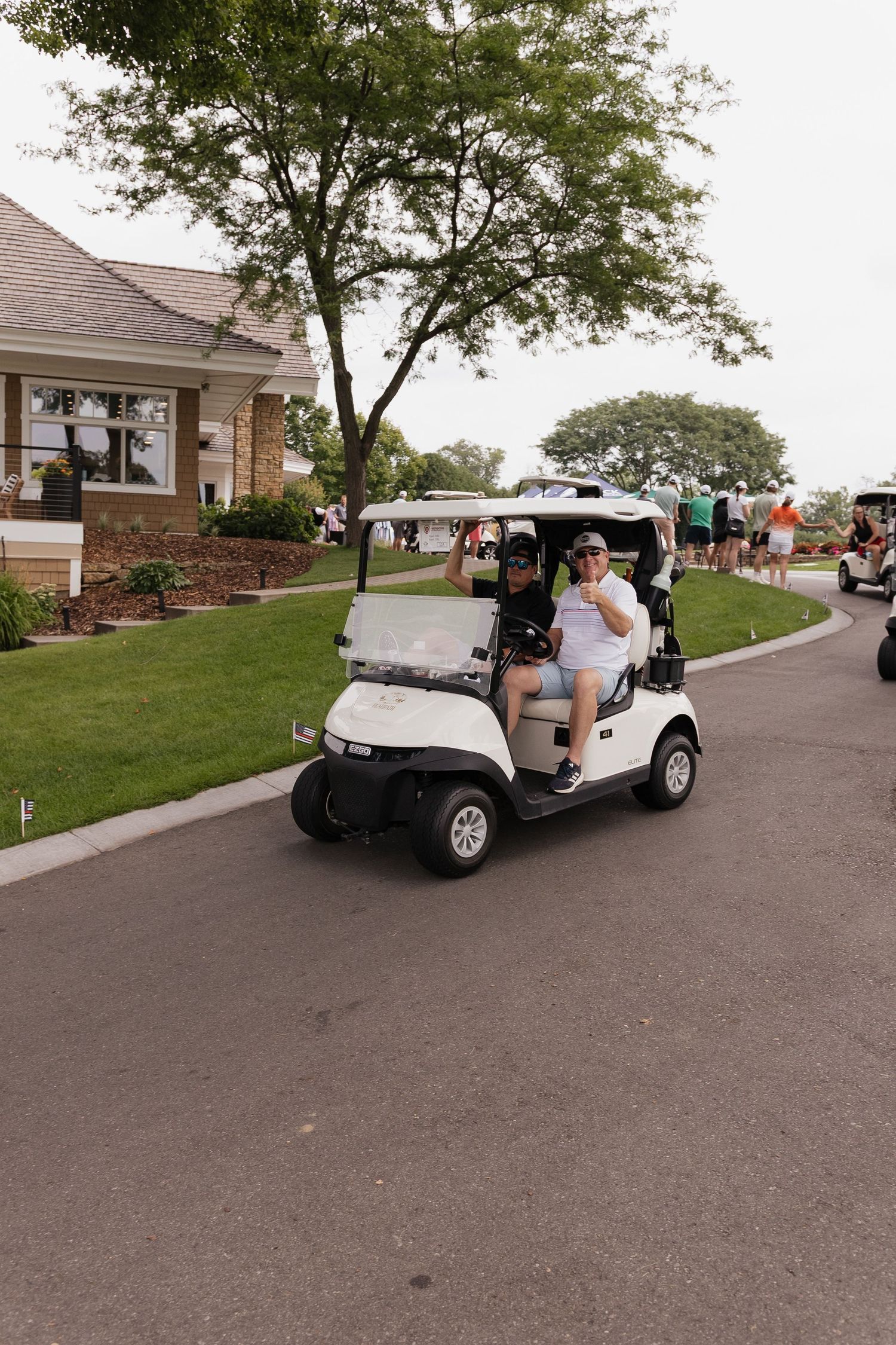 Two smiling golfers wave from a white golf cart on a sunny golf course path during a tournament.