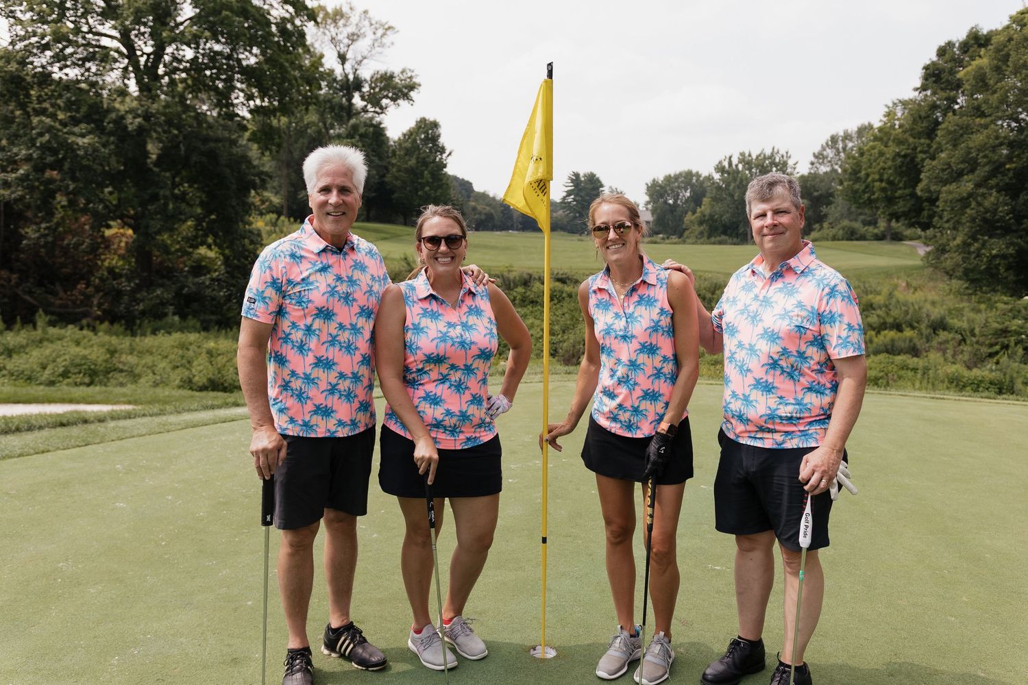 Four golfers in matching floral polo shirts pose together on a golf green beside a yellow flag.