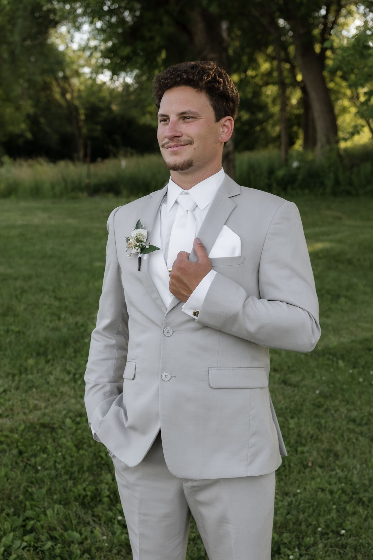 Groom posing confidently in a light gray suit with white boutonniere and pocket square in an outdoor setting.