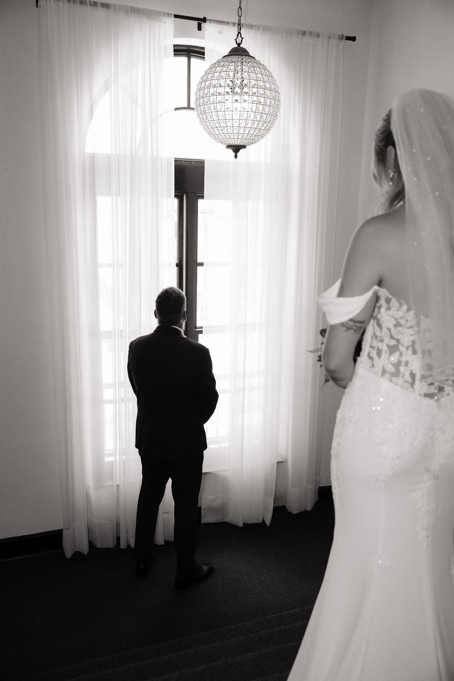Black and white photo of a bride in lace gown approaching groom standing at window before first look.