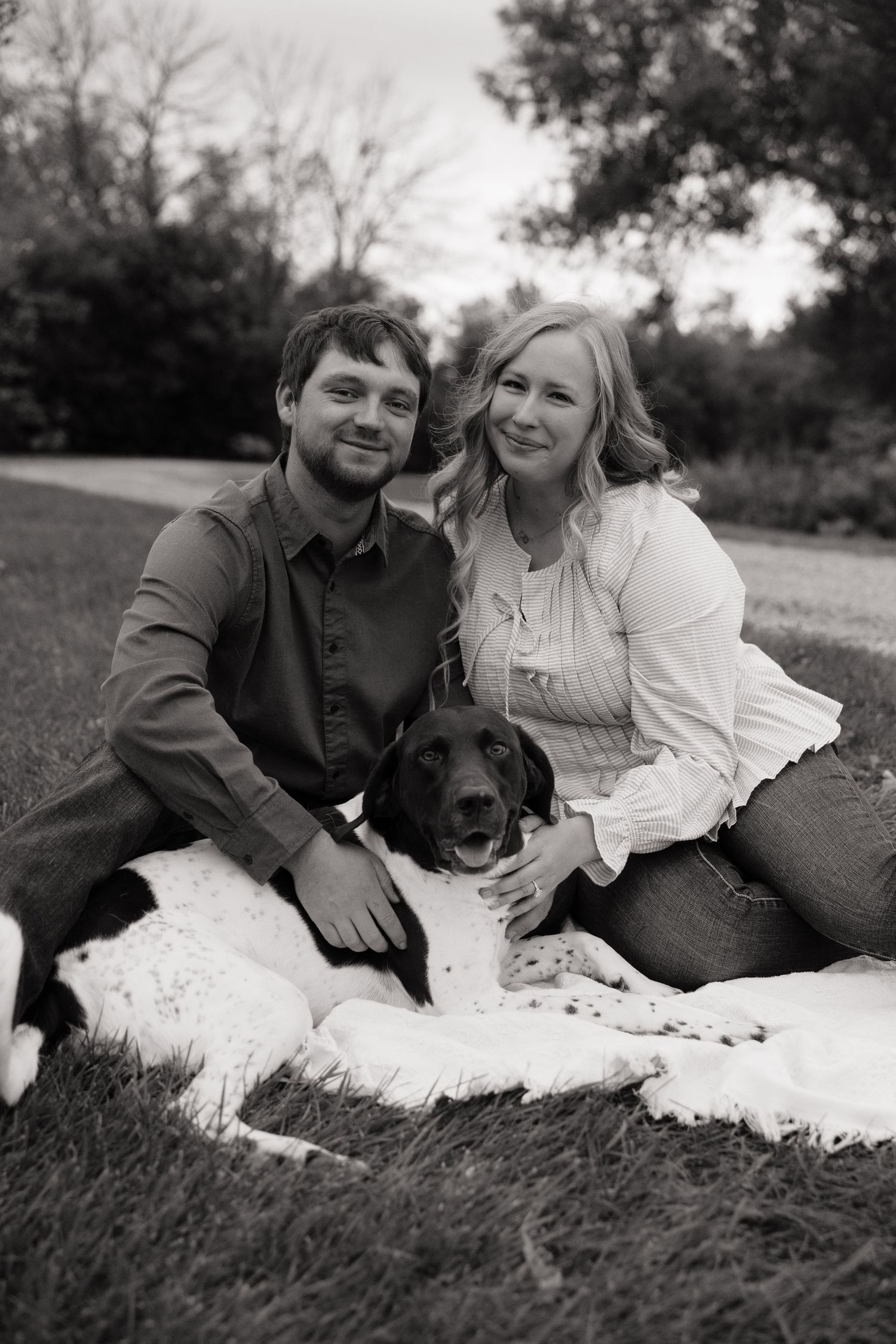 Black and white photo of a smiling couple sitting on a blanket in a park with their happy dog.
