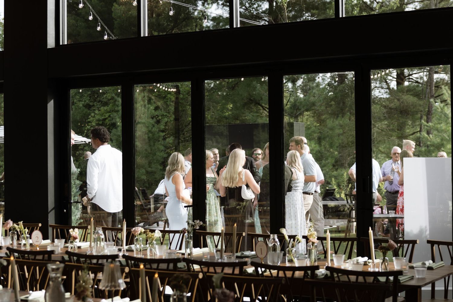 Elegant wedding reception venue with long dining tables set with floral arrangements and guests mingling near floor-to-ceiling windows.