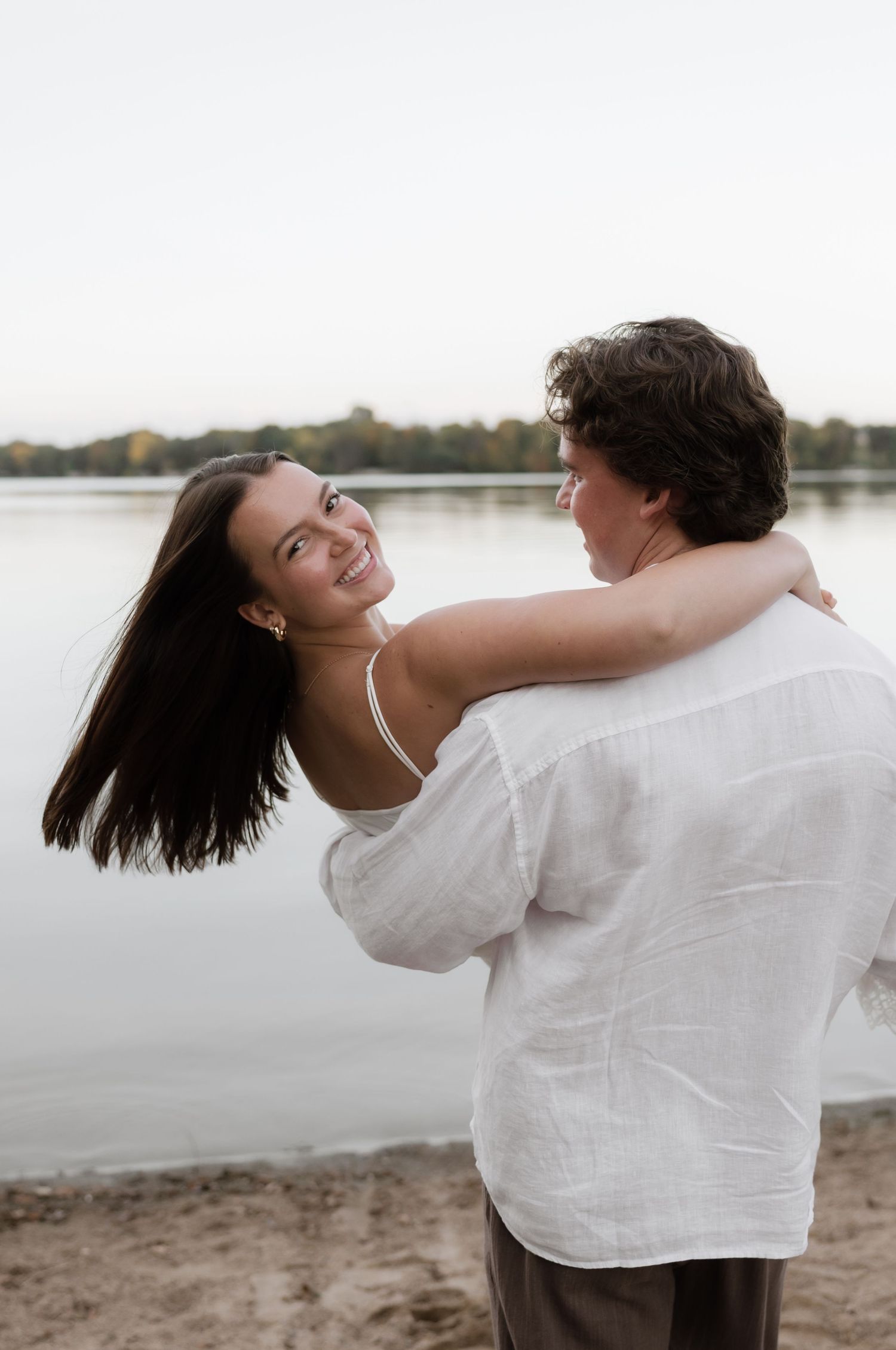 Man lifting and spinning woman in white dress on sandy lakeside beach at dusk, both smiling.