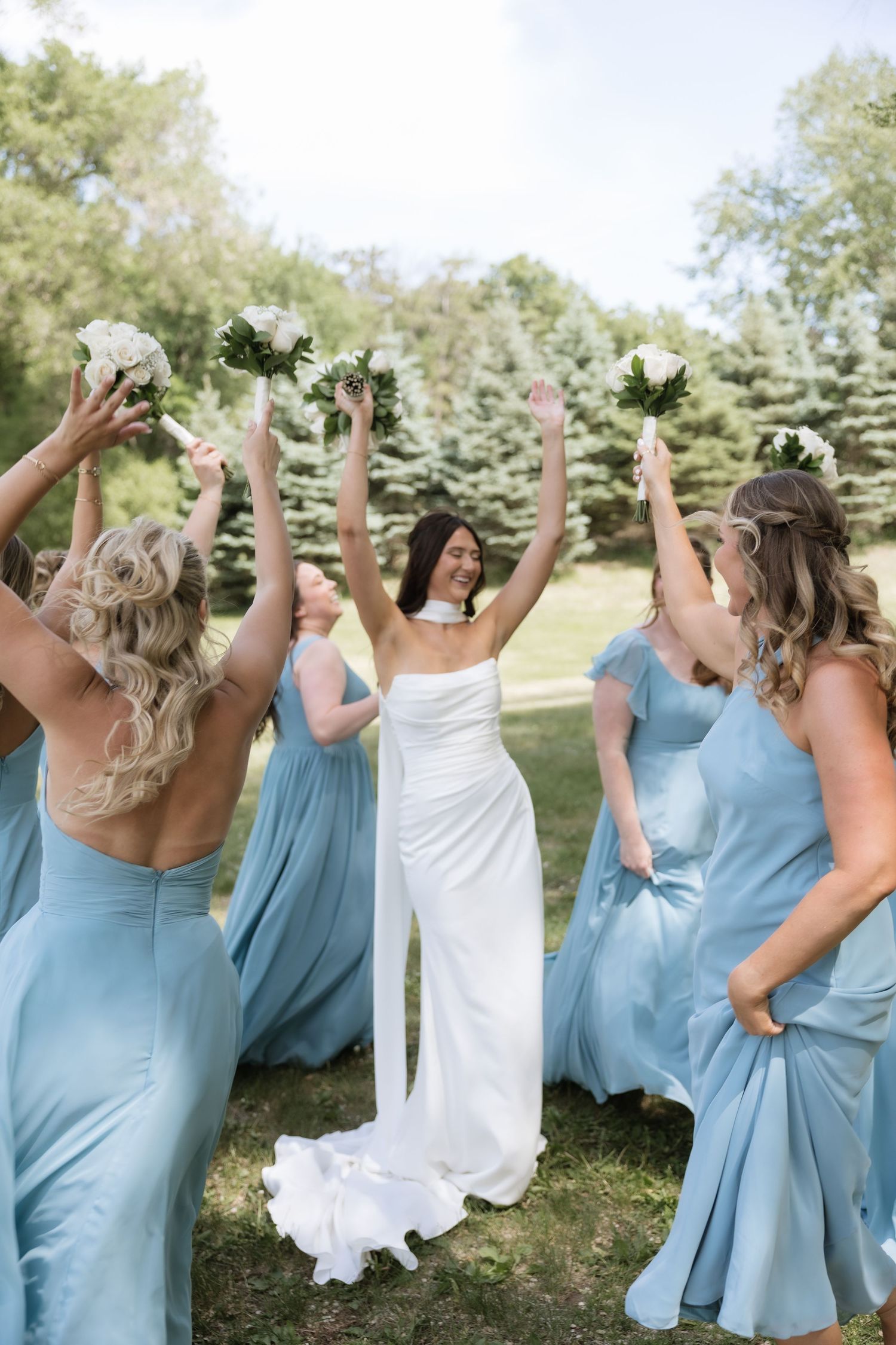 Bride in white halter gown celebrates with arms raised surrounded by bridesmaids in dusty blue dresses.