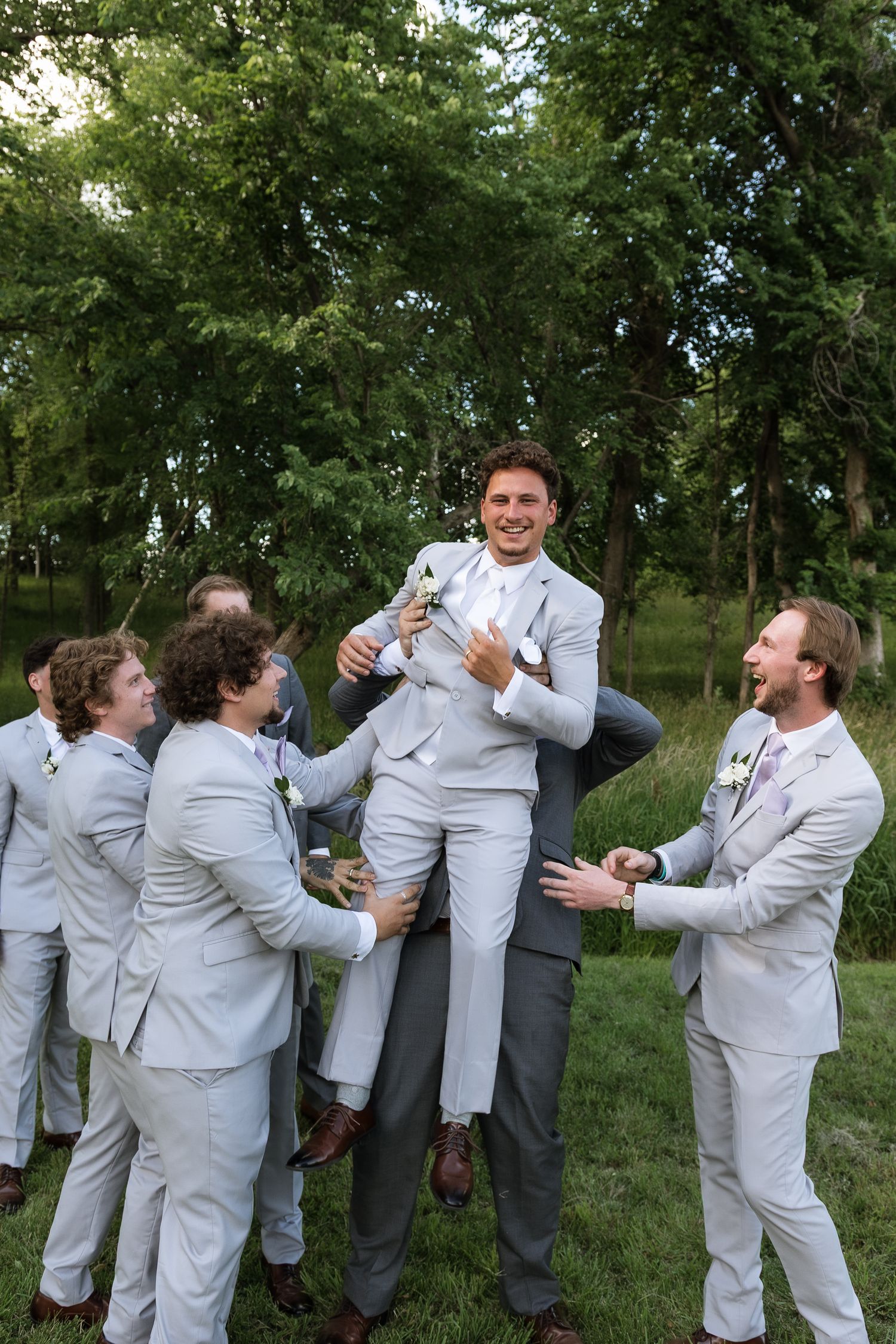 Groomsmen in light gray suits joyfully lifting the groom outdoors with lush green trees in the background.