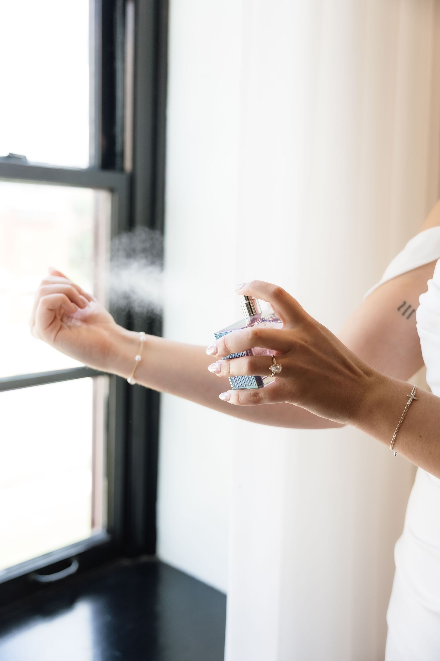 A bride sprays perfume on her wrist near a window, wearing bracelets and a ring on her wedding day.