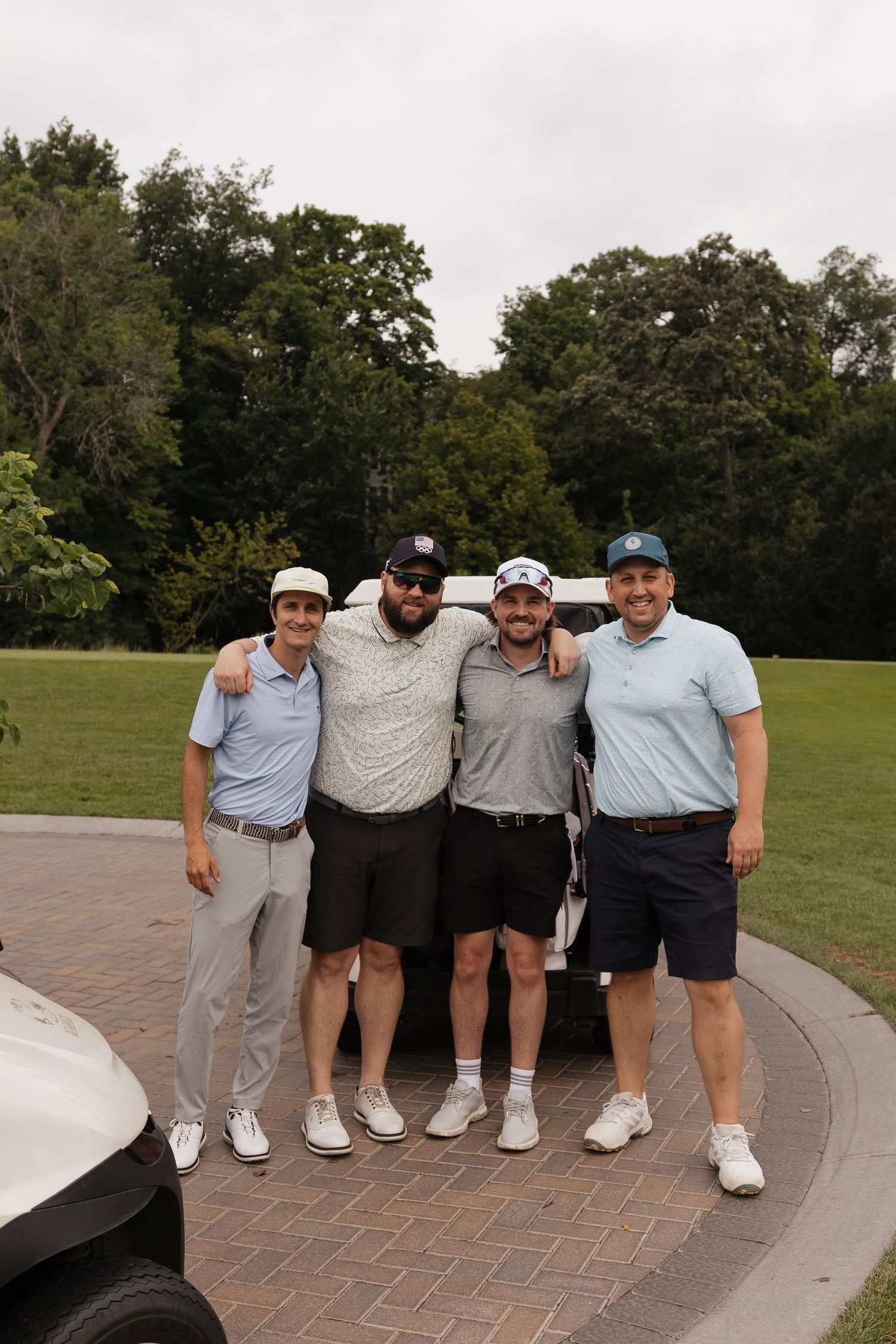 Four men in golf attire smiling together on a lush green golf course near a cart path.