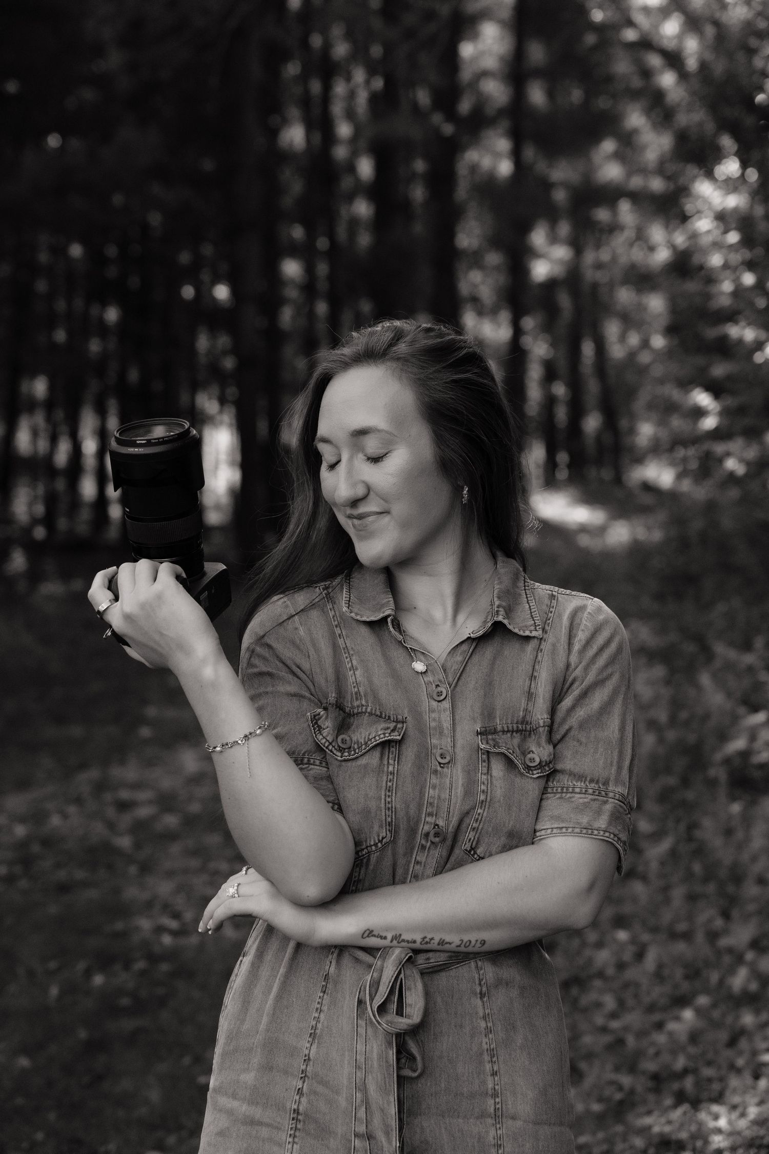 Black and white photo of a woman in a denim dress holding a camera in a forest setting.