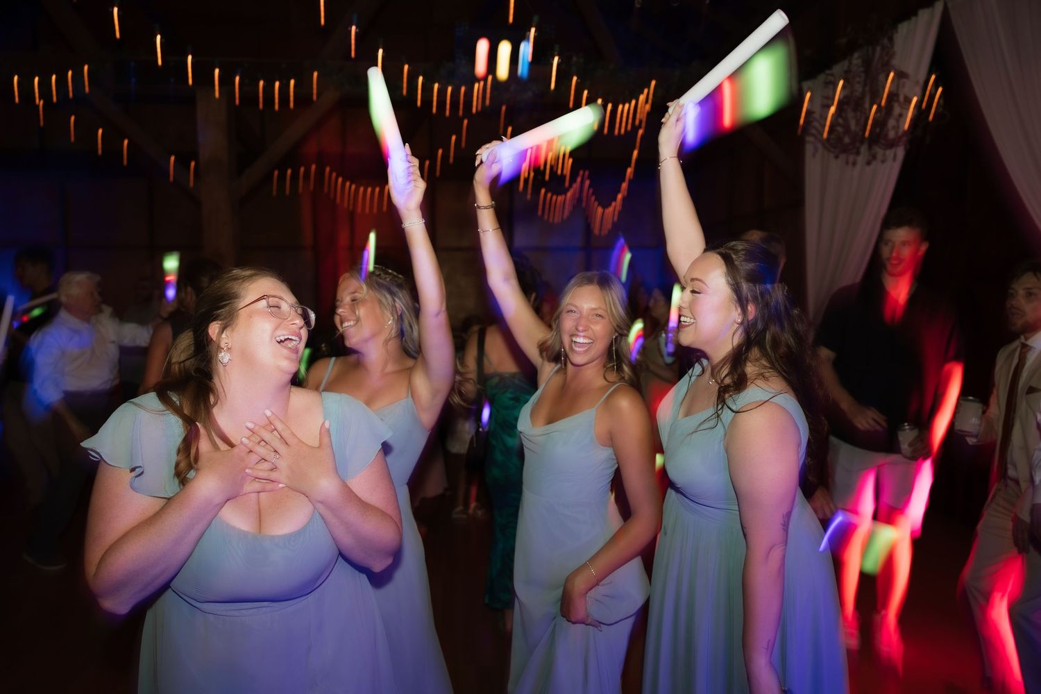 Bridesmaids in light blue dresses dance and wave glow sticks joyfully at a vibrant wedding reception party.