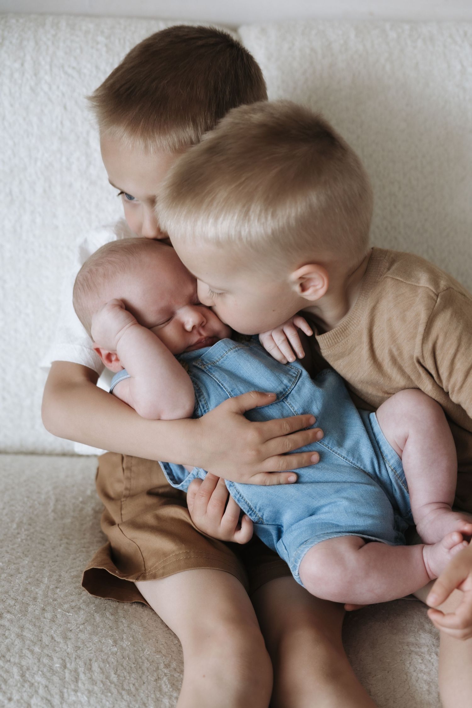 Two older brothers lovingly hold and kiss their sleeping newborn sibling dressed in a blue romper.