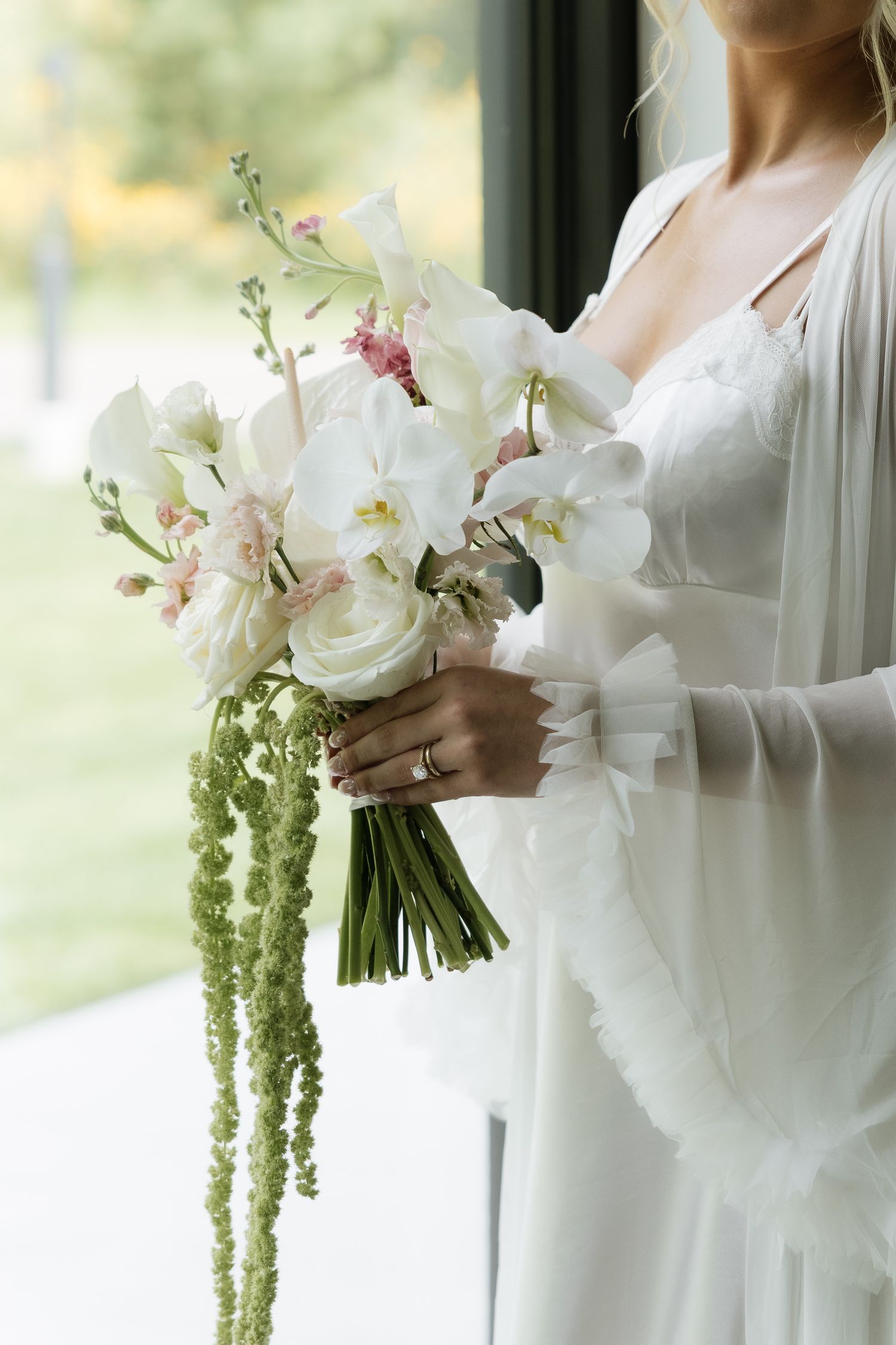 Bride in white gown holding elegant bouquet of white orchids, roses, and trailing green amaranthus near a window.