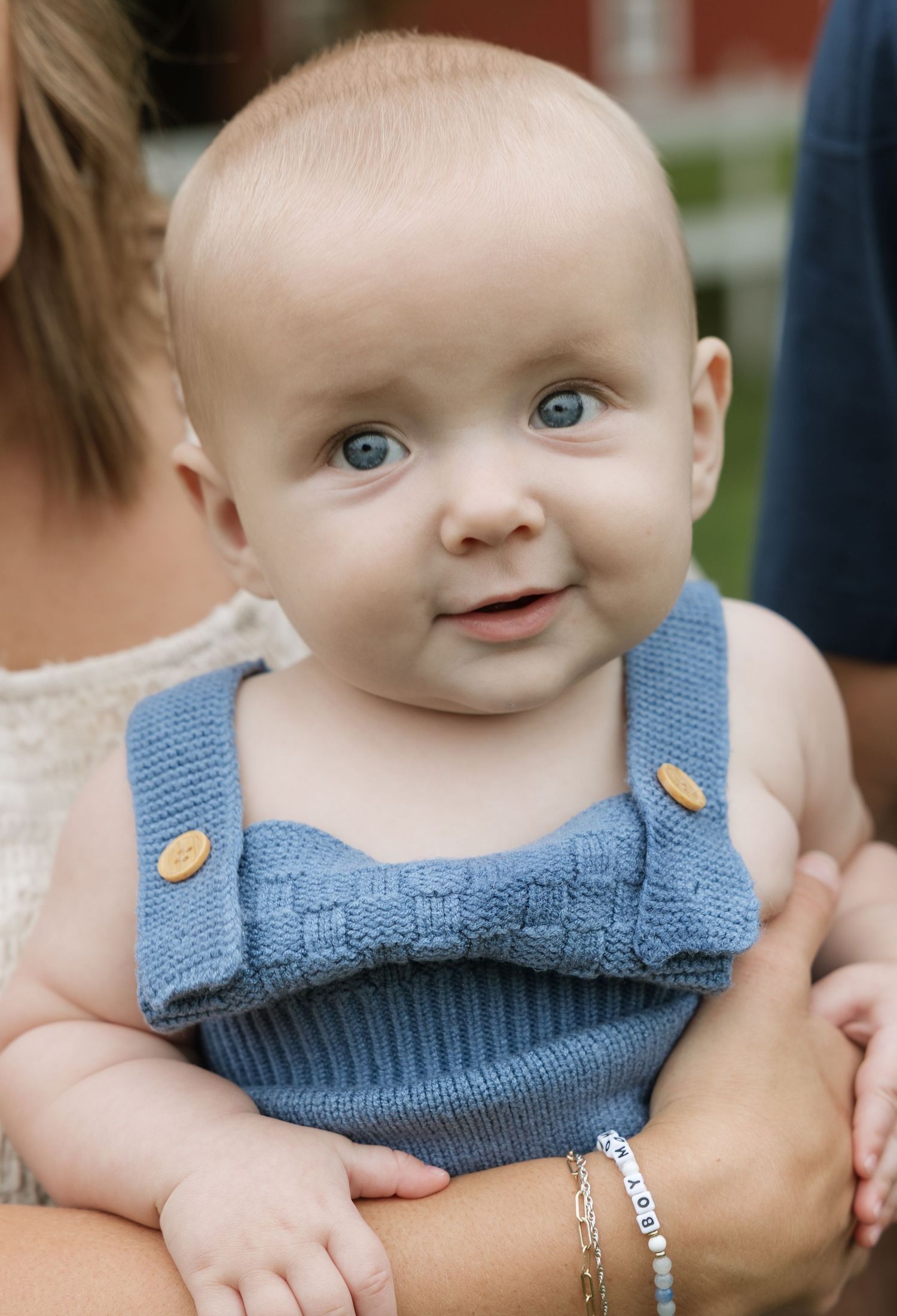 Smiling baby with blue eyes wearing a blue knitted romper being held by an adult outdoors.