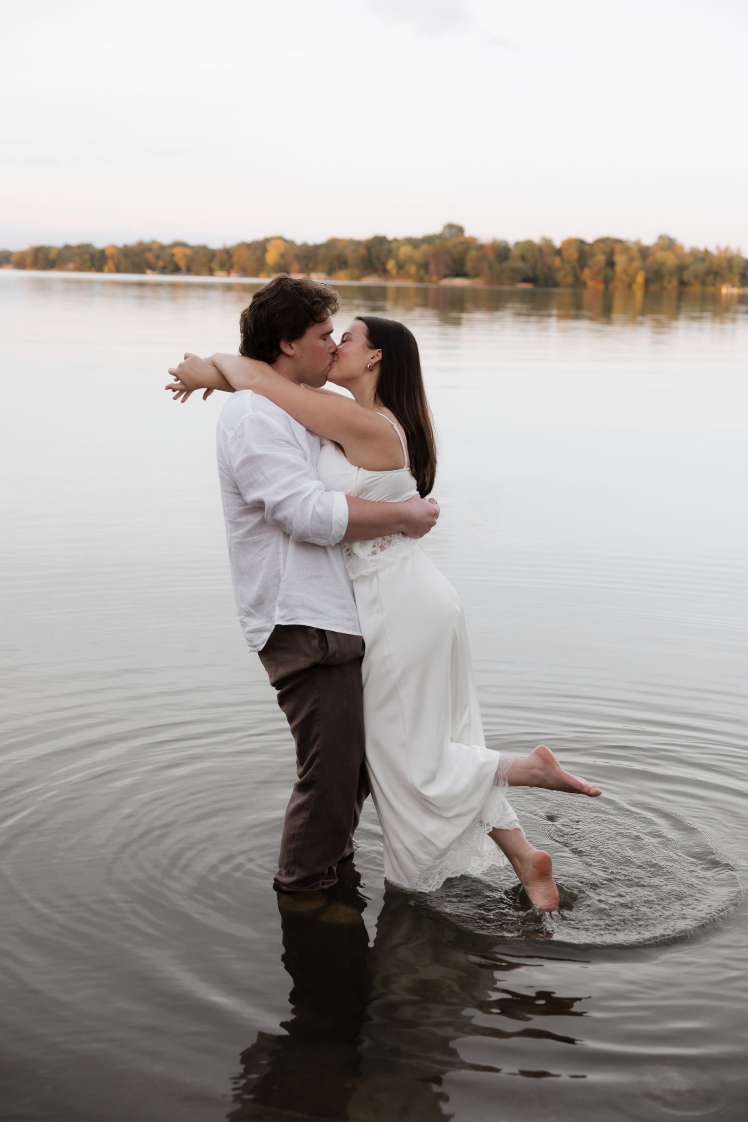 Couple kissing romantically while standing in a lake, woman in white dress lifted by man in white shirt.