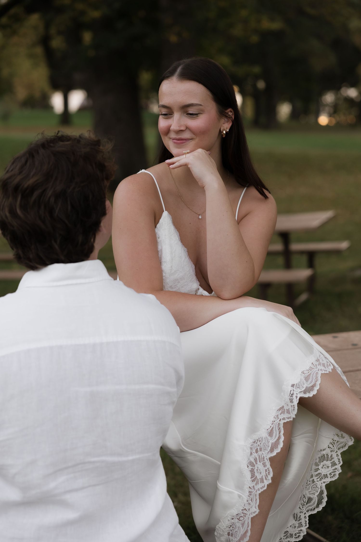 Woman in white lace dress sitting on table smiling softly at man in white shirt in park at dusk.