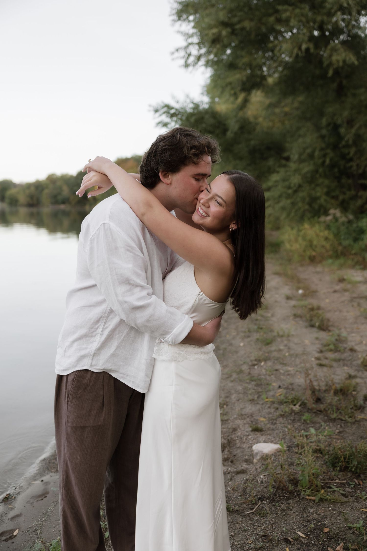 A couple embraces romantically by a lakeside, the woman in a white dress smiling joyfully as the man kisses her cheek.