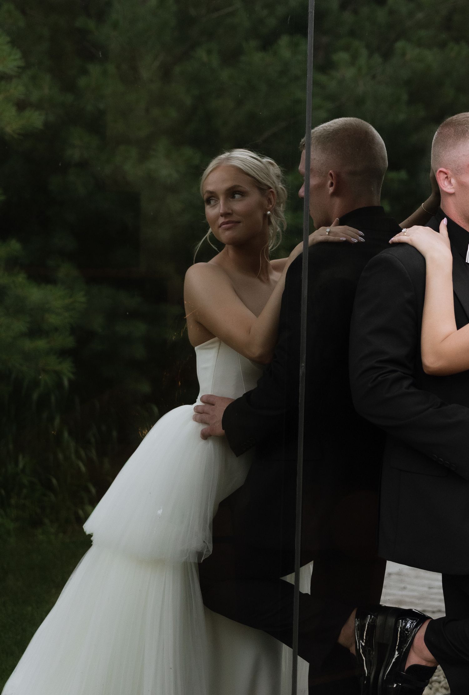Bride in white gown embraces groom in black suit outdoors, reflected in mirror, with lush green trees in background.