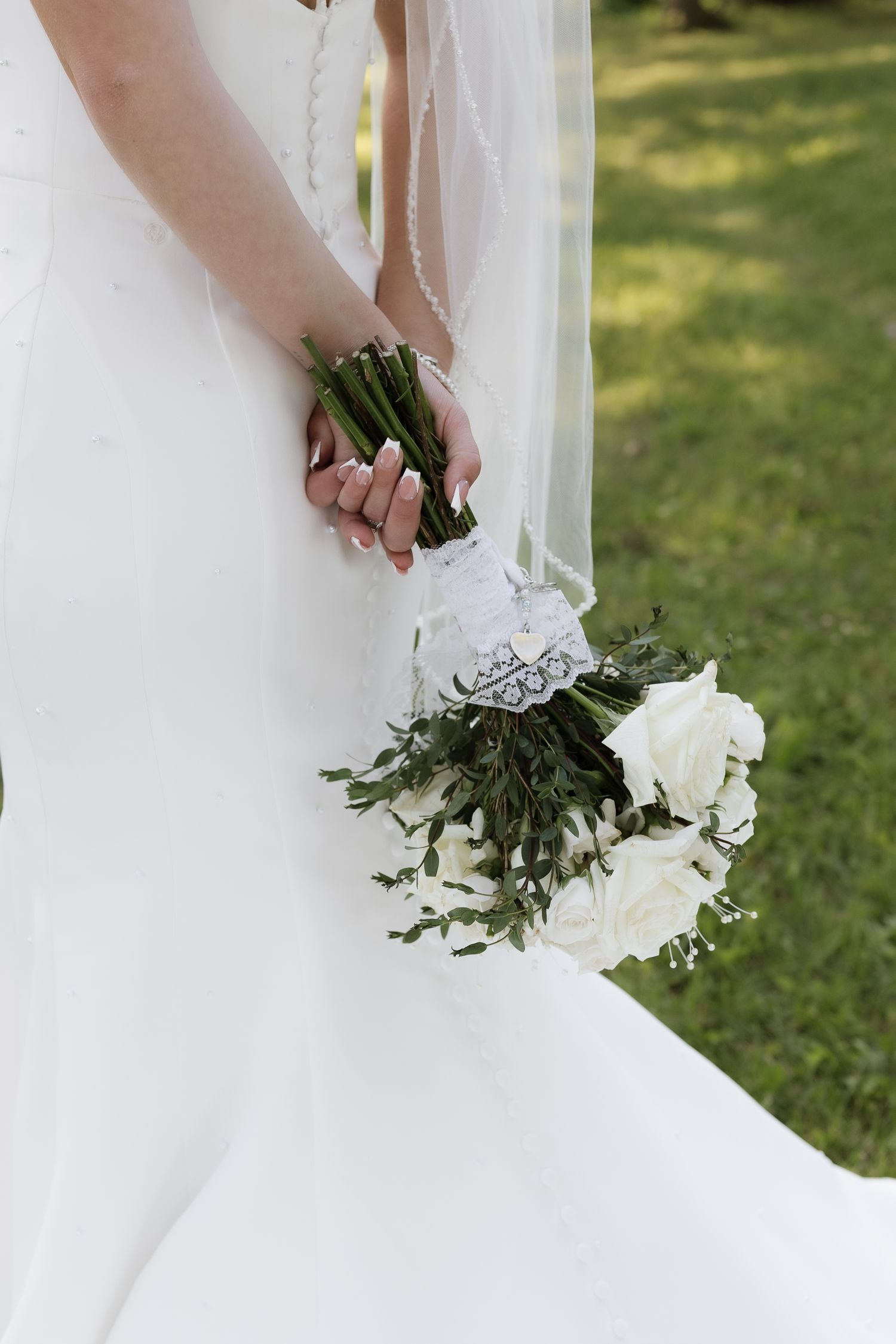 Bride in white gown holding elegant bouquet of white roses with greenery, tied with lace ribbon, outdoors.
