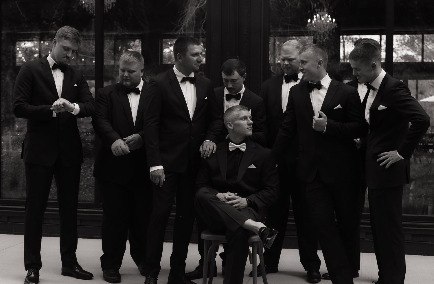 Black and white photo of groomsmen in tuxedos gathered around seated groom at elegant wedding venue.