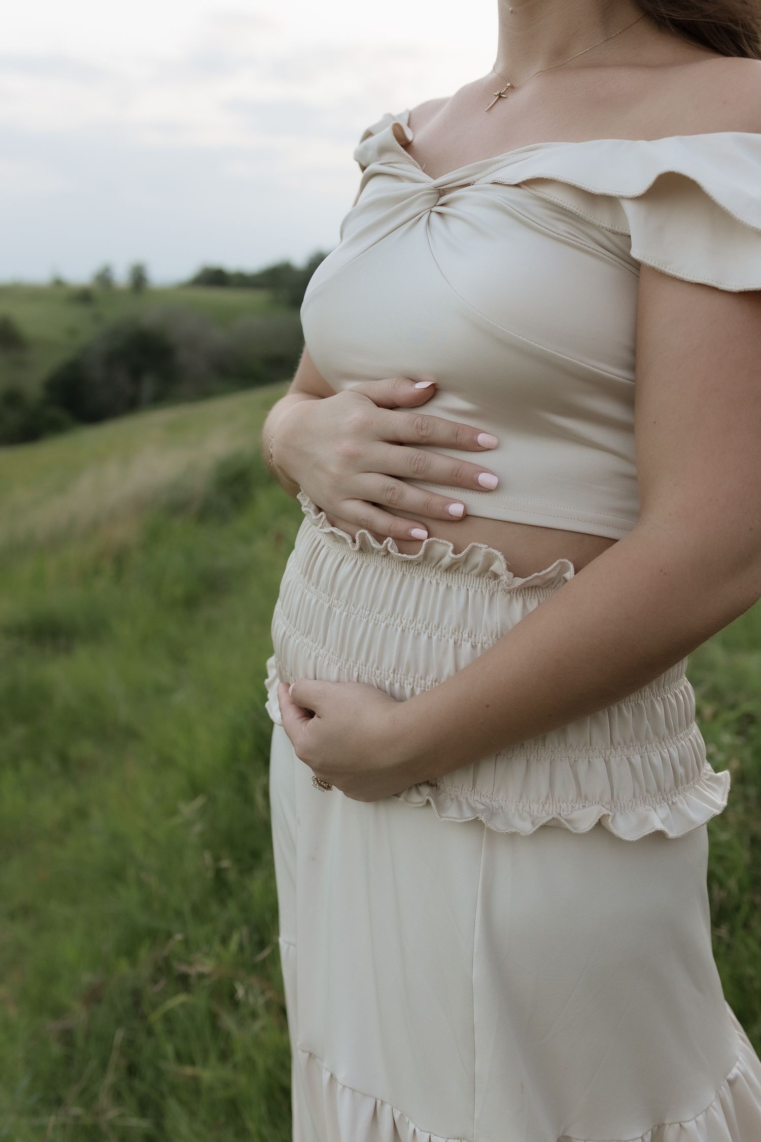 Pregnant woman in cream off-shoulder crop top and smocked skirt cradling baby bump in green field.