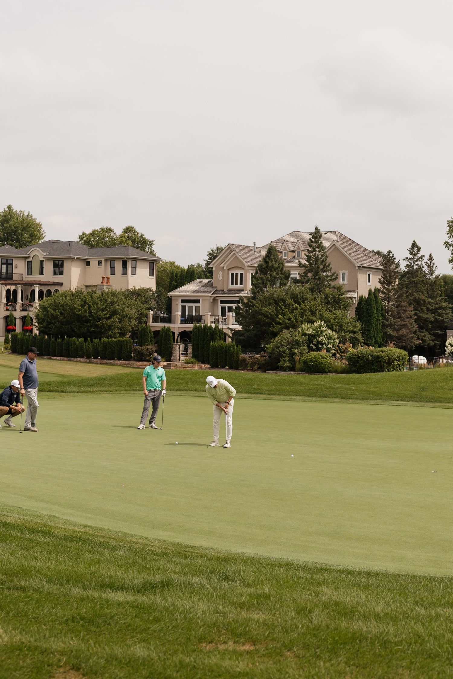 Golfers on a lush green putting course surrounded by elegant luxury homes and manicured trees on a cloudy day.