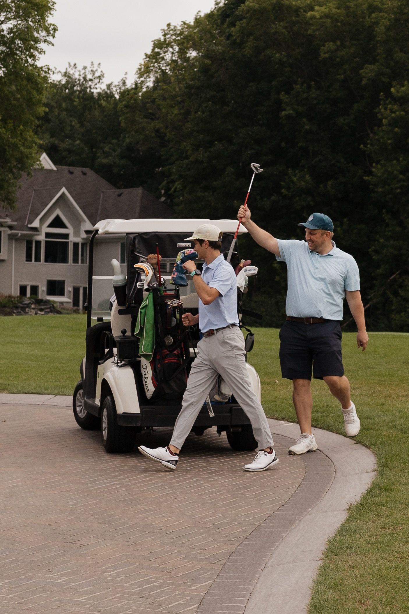 Two golfers walk beside a golf cart on a course path, one raising a club, with trees and a house in the background.