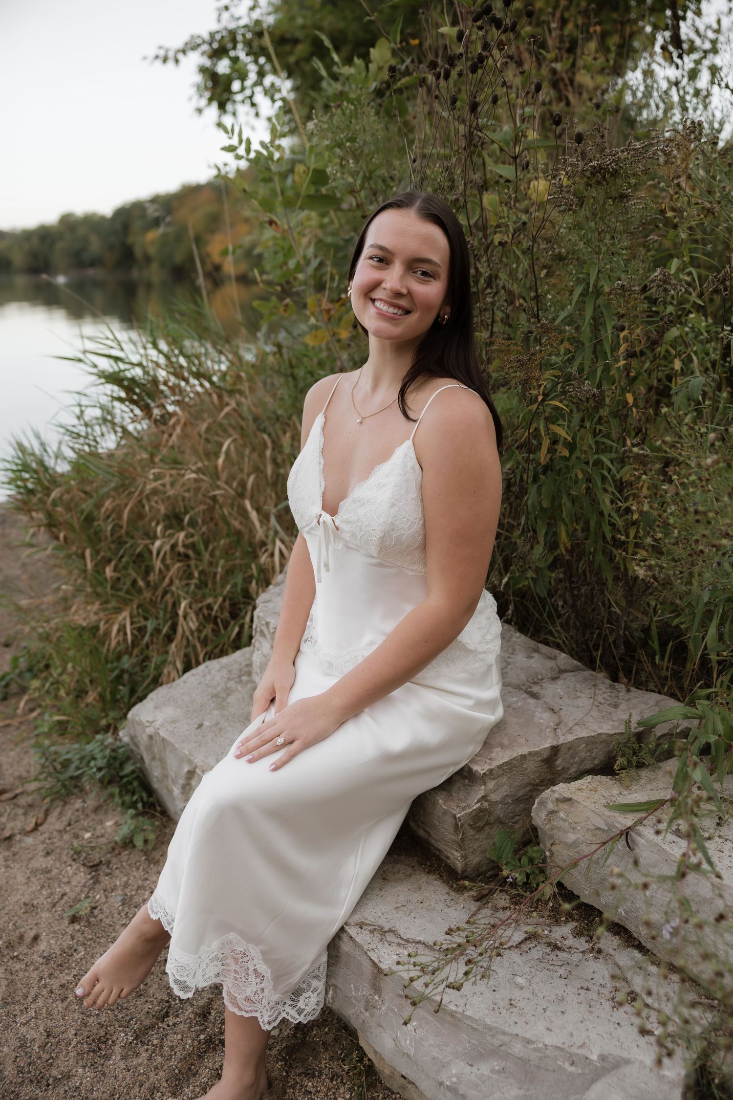 Smiling brunette woman in white lace slip dress sitting on rocks by a lakeside surrounded by greenery.