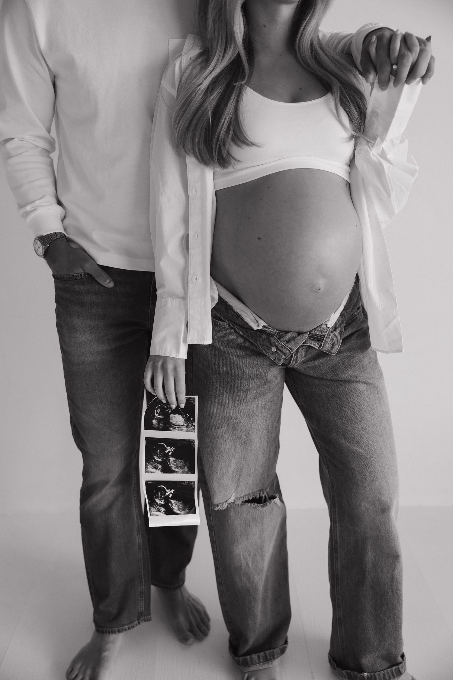 Black and white photo of couple holding ultrasound images beside pregnant belly in white shirts and jeans.