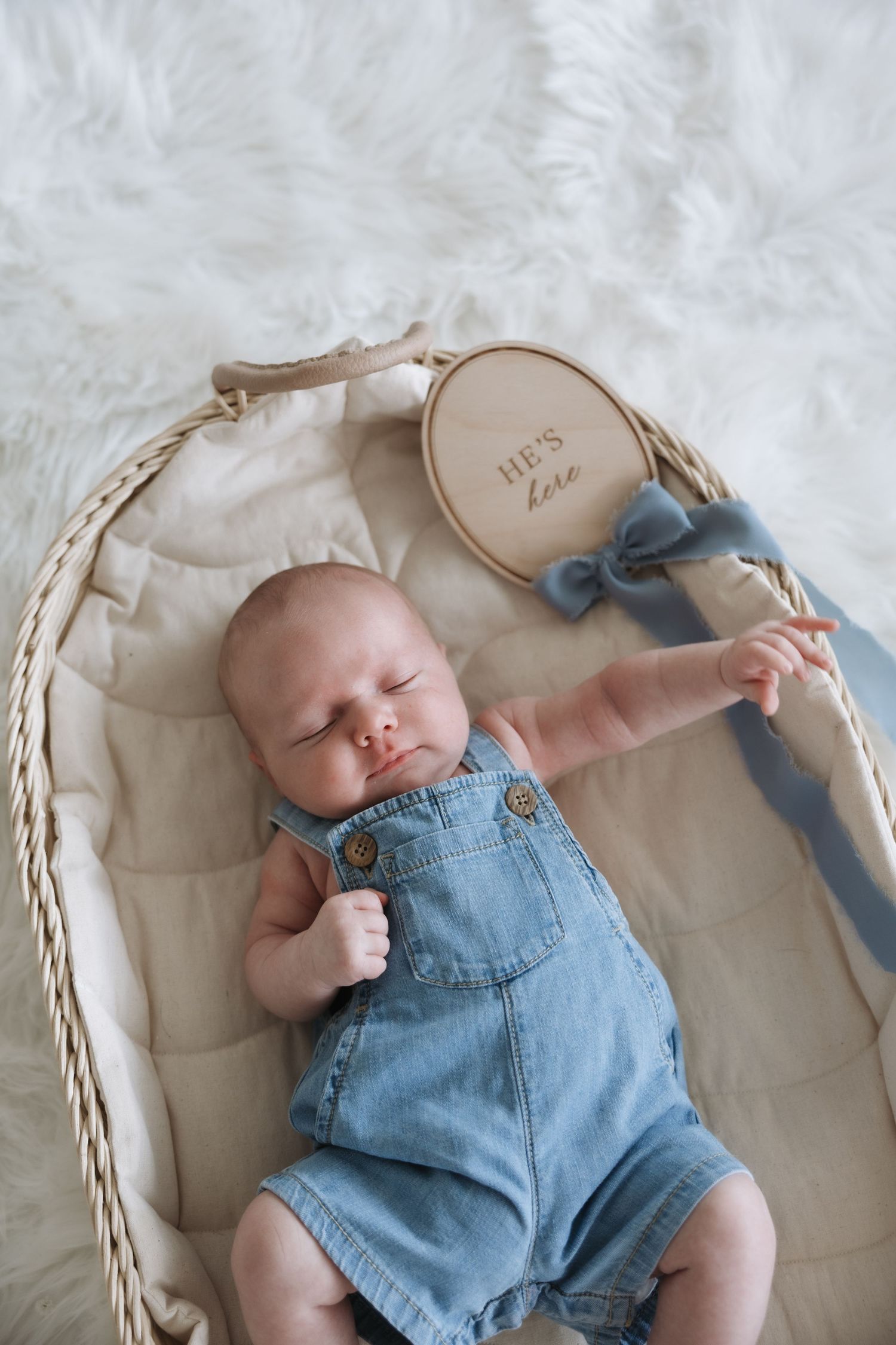 Newborn baby in denim overalls resting in wicker basket with he's here wooden milestone sign.