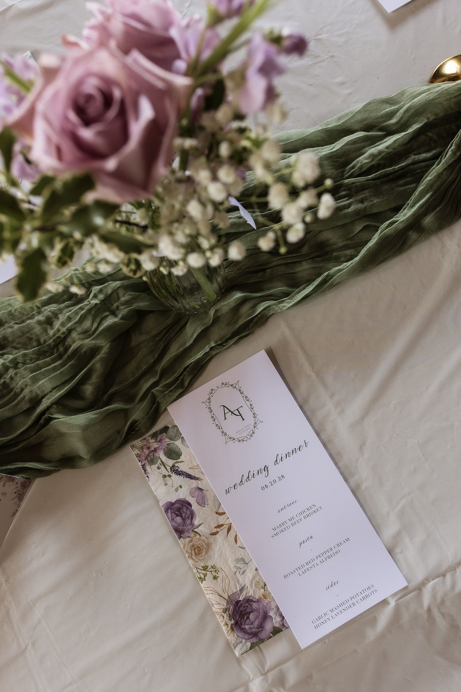Elegant wedding dinner menu with floral details beside lavender roses and green cheesecloth table runner.