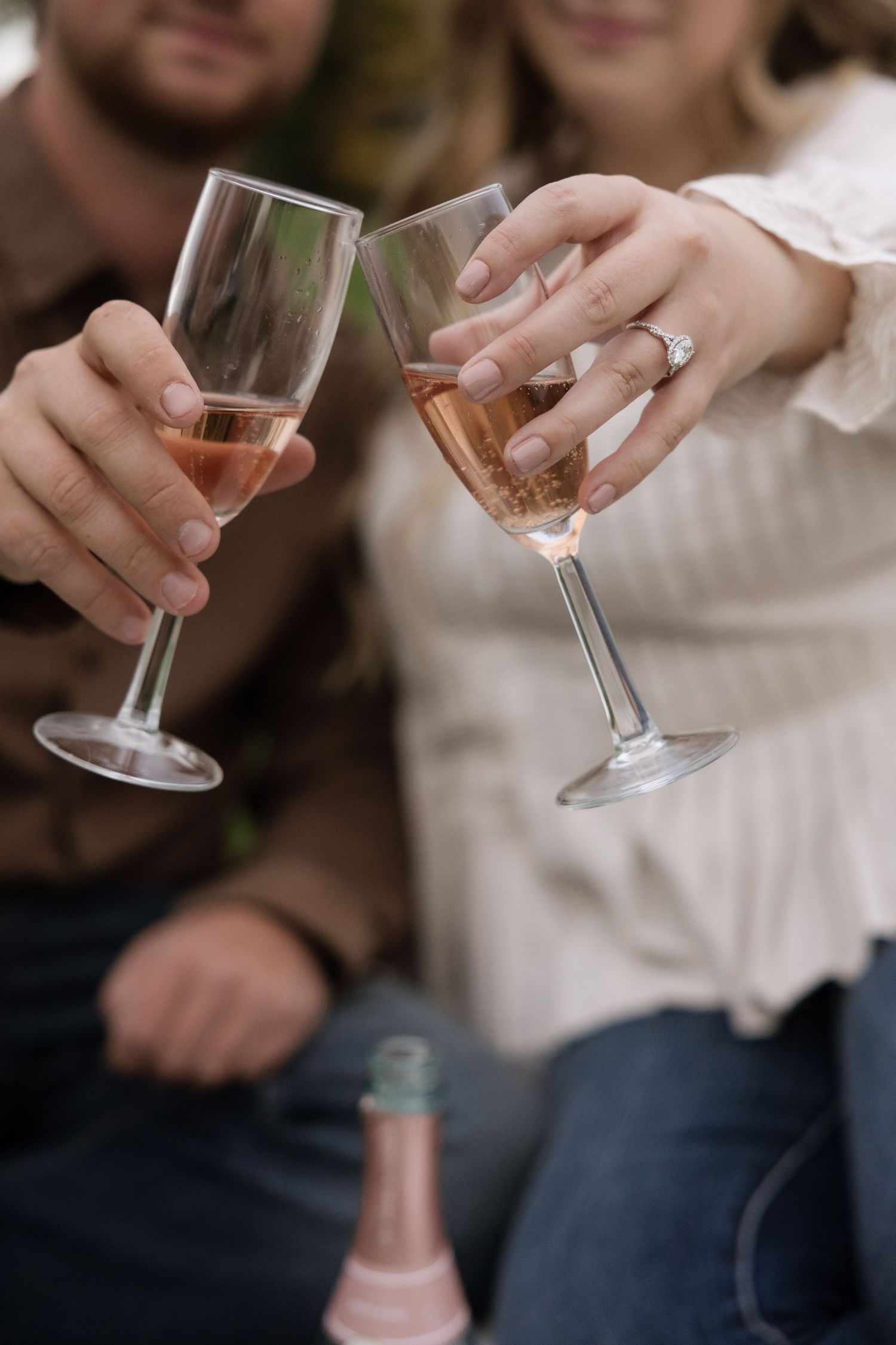 Couple toasting with rose wine glasses, woman wearing engagement ring, celebration outdoors.