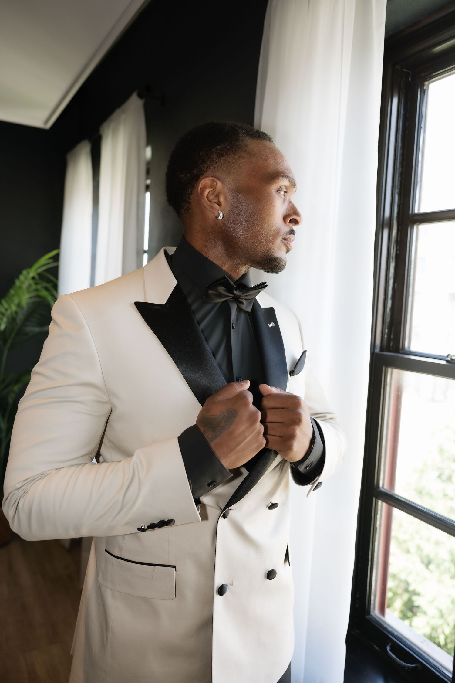 A stylish man in a white tuxedo jacket and black bow tie adjusts his lapel while standing by a window.