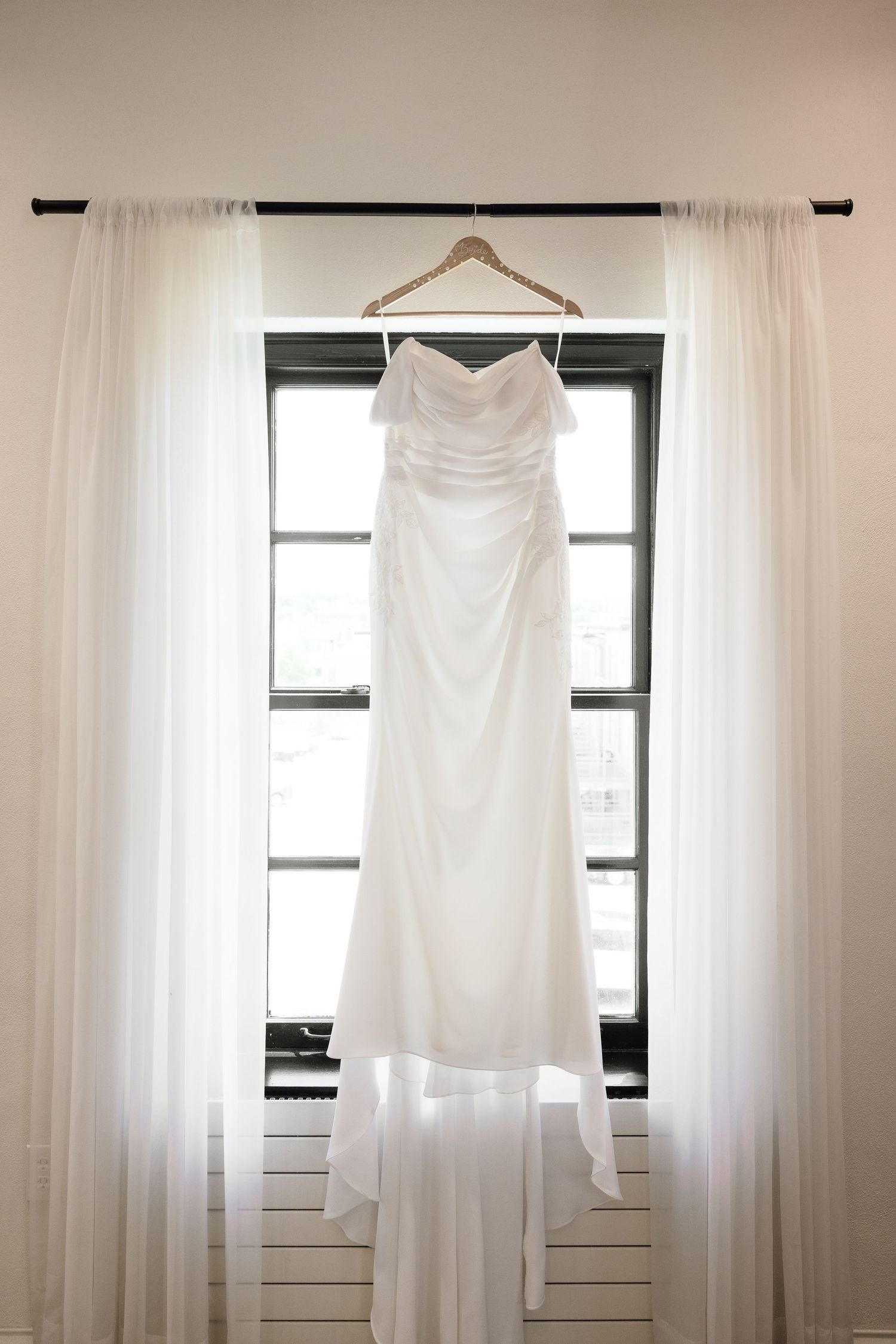 Elegant strapless white wedding gown hanging on a wooden hanger in front of a bright window with sheer curtains.