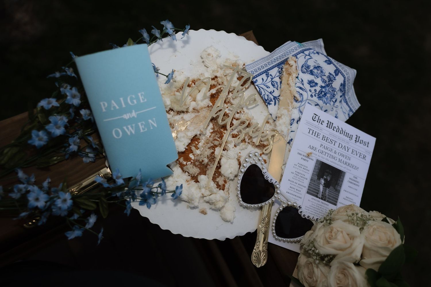 Wedding flat lay with Paige and Owen program, white floral cake, heart sunglasses, and newspaper announcement.