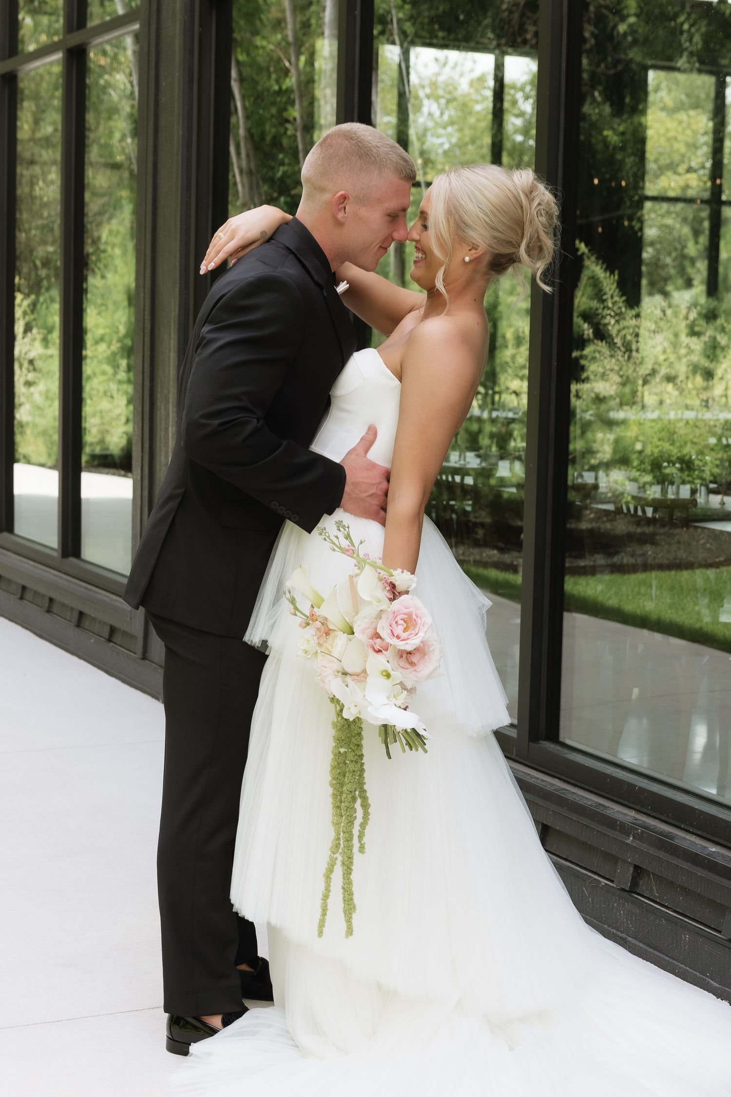 Bride and groom share romantic moment holding cascading pink bouquet before large glass windows.