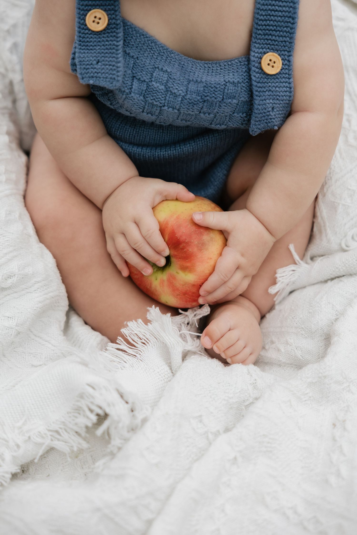 Baby in blue knit overalls holding a red and yellow apple while sitting on a white blanket.