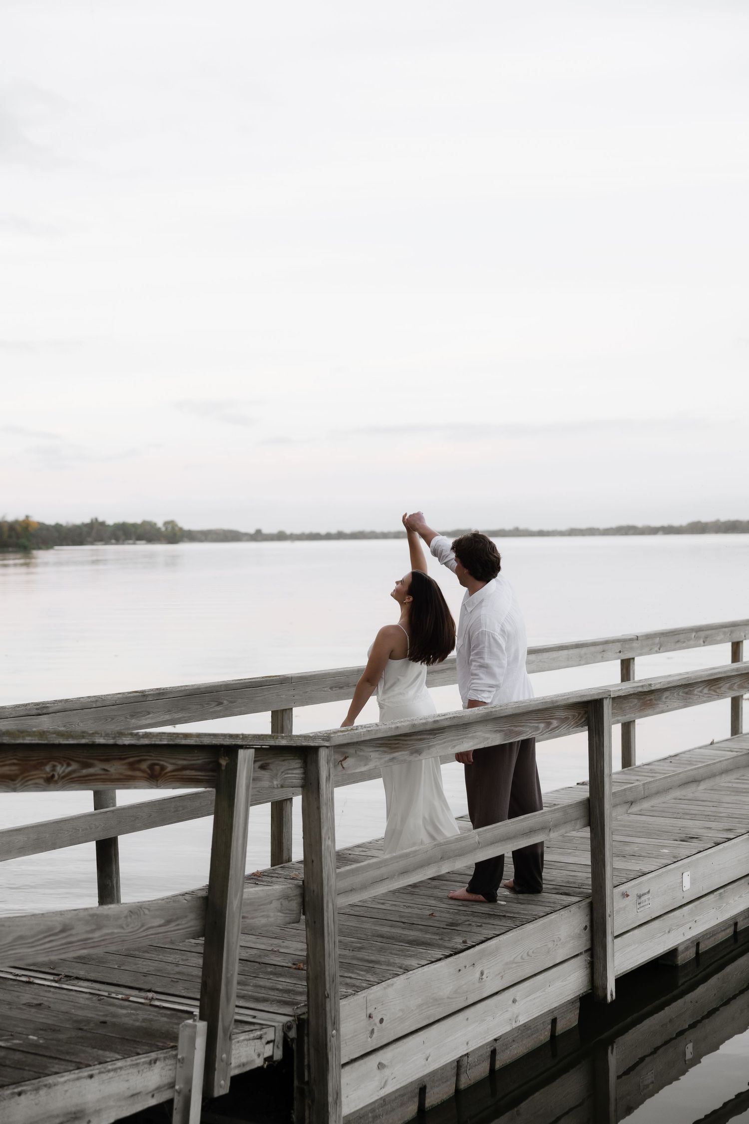 Engaged couple sitting on a wooden dock over a serene lake, man spinning woman's hand in the air.