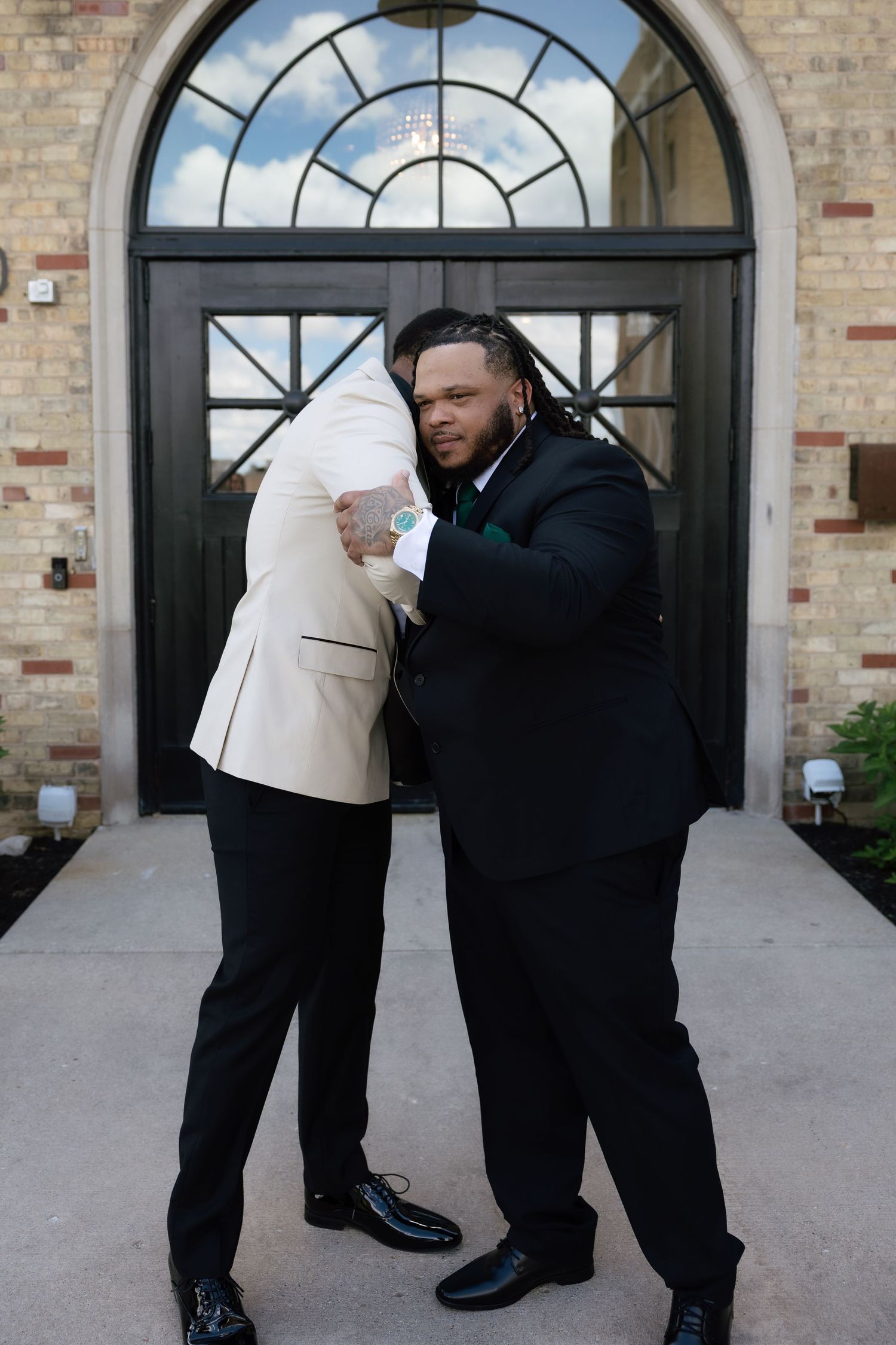 Two grooms in formal suits share a heartfelt hug outside an elegant brick building with arched doors.