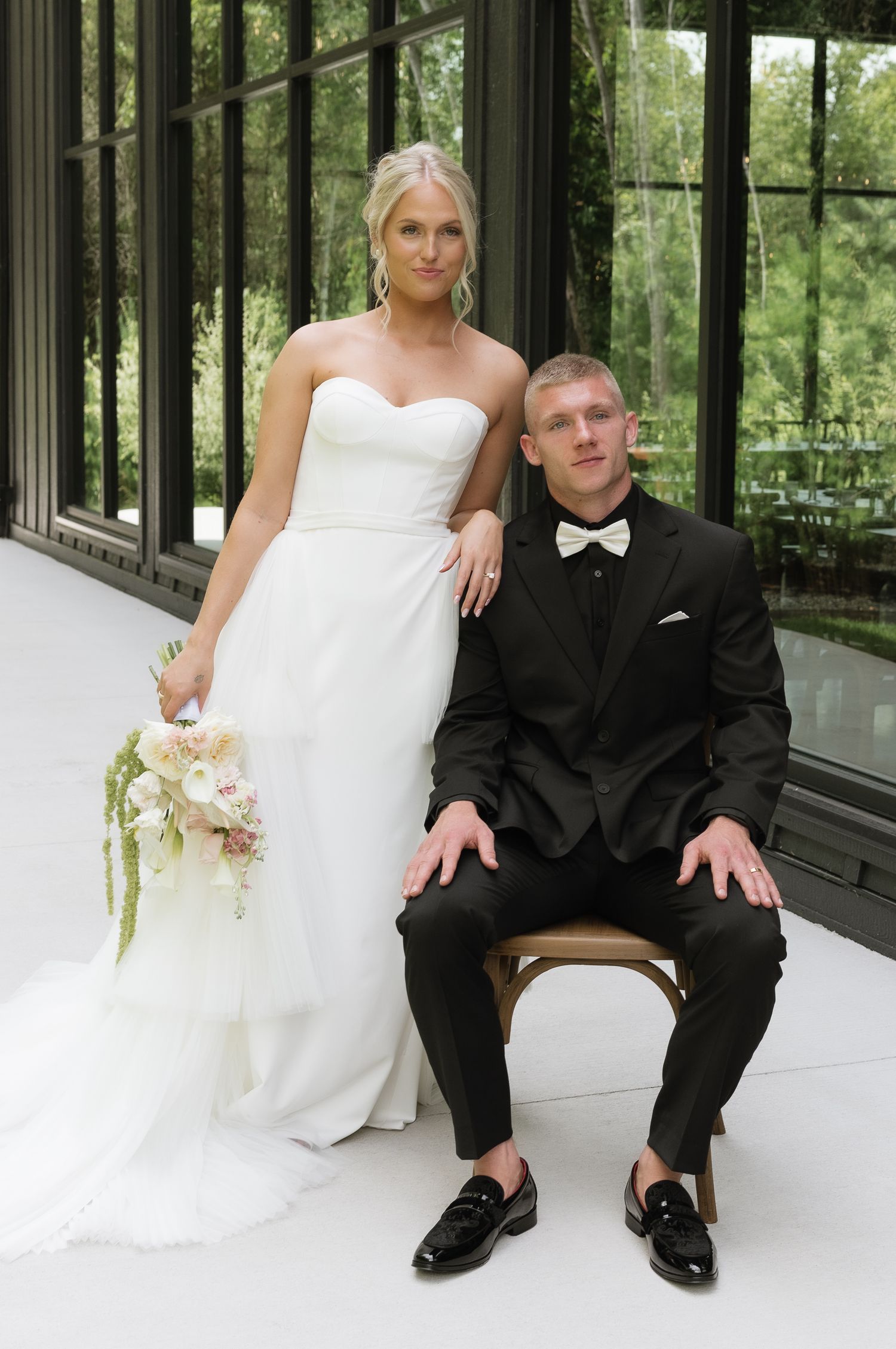 Bride stands beside seated groom in black tuxedo with bow tie inside glass-walled venue.
