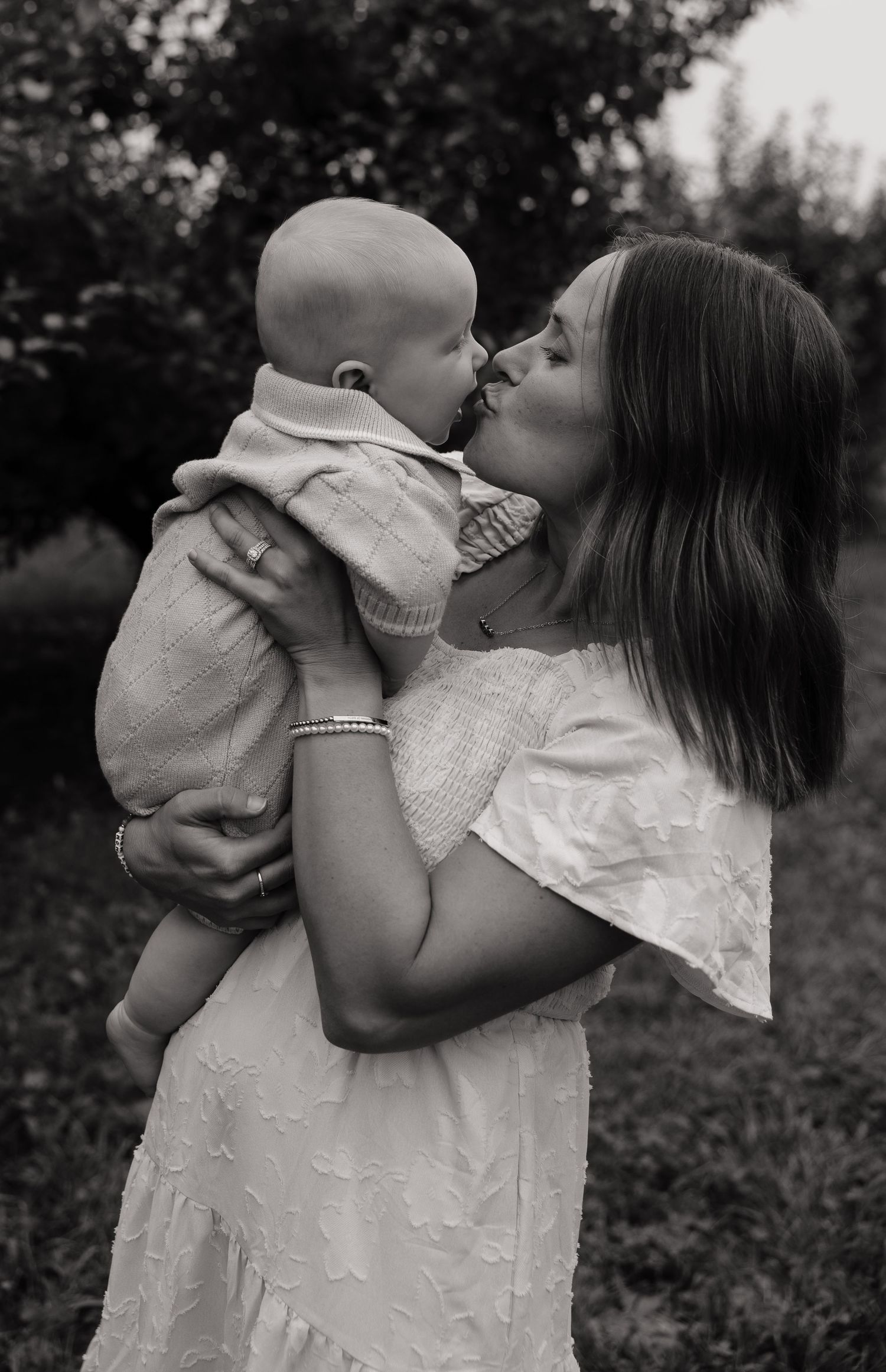 Black and white photo of a mother lovingly kissing her baby while holding him up in an outdoor garden setting.