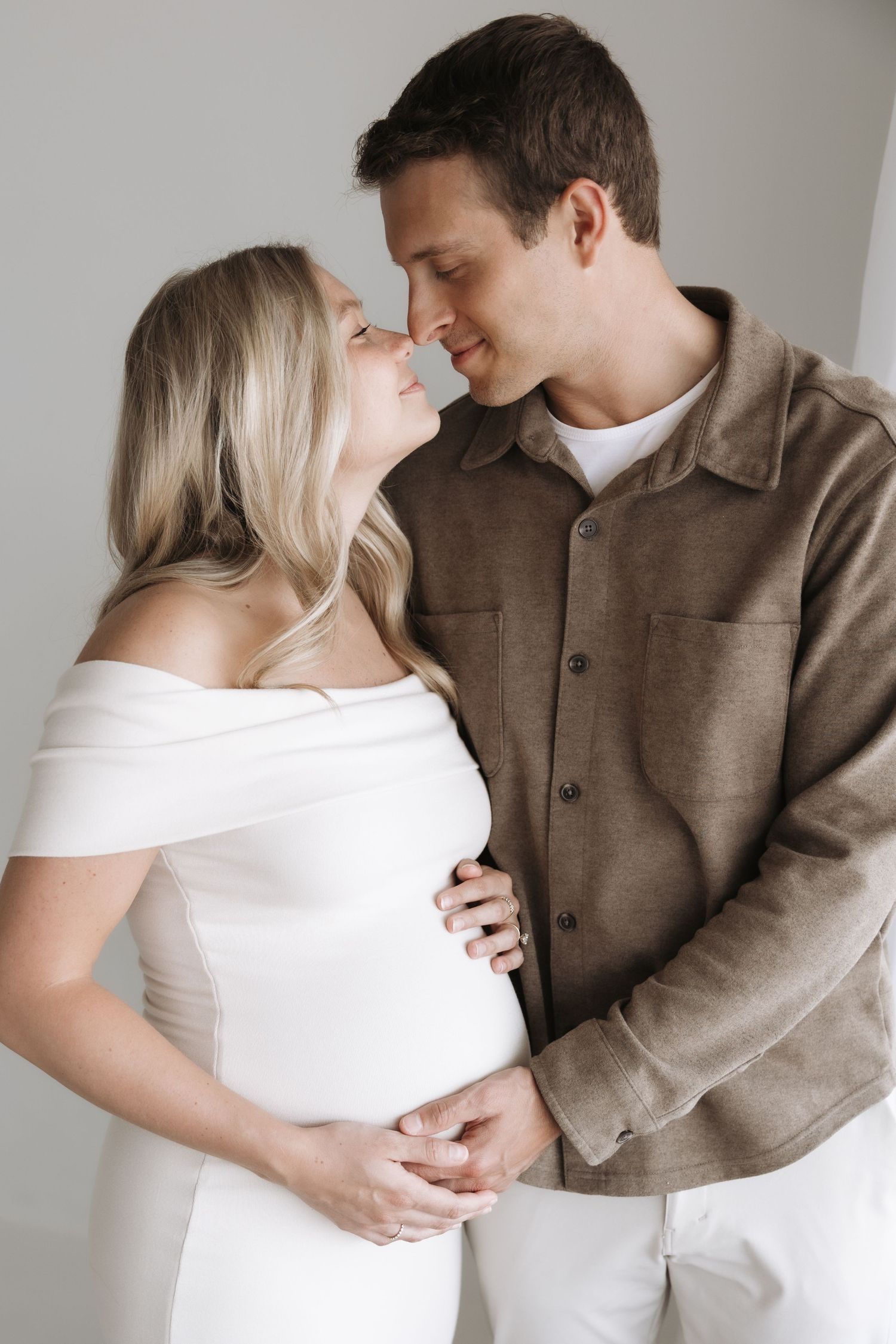 A couple shares a tender moment, the pregnant woman in white dress held by her partner in a brown shirt.