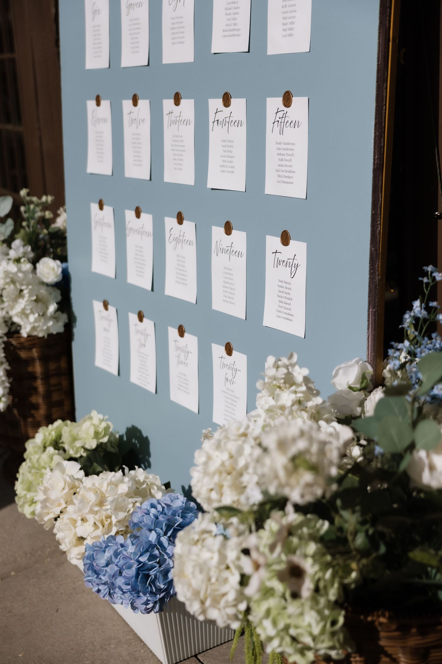 Blue wedding seating chart board with white cards and gold pins, surrounded by white and blue hydrangea floral arrangements.
