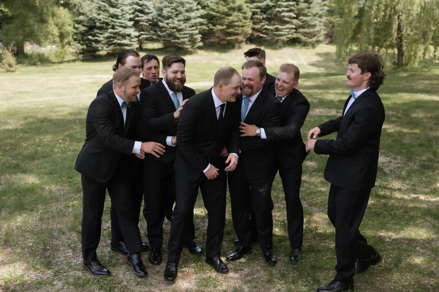 Groom surrounded by groomsmen in black suits sharing laughs on green lawn during wedding day.