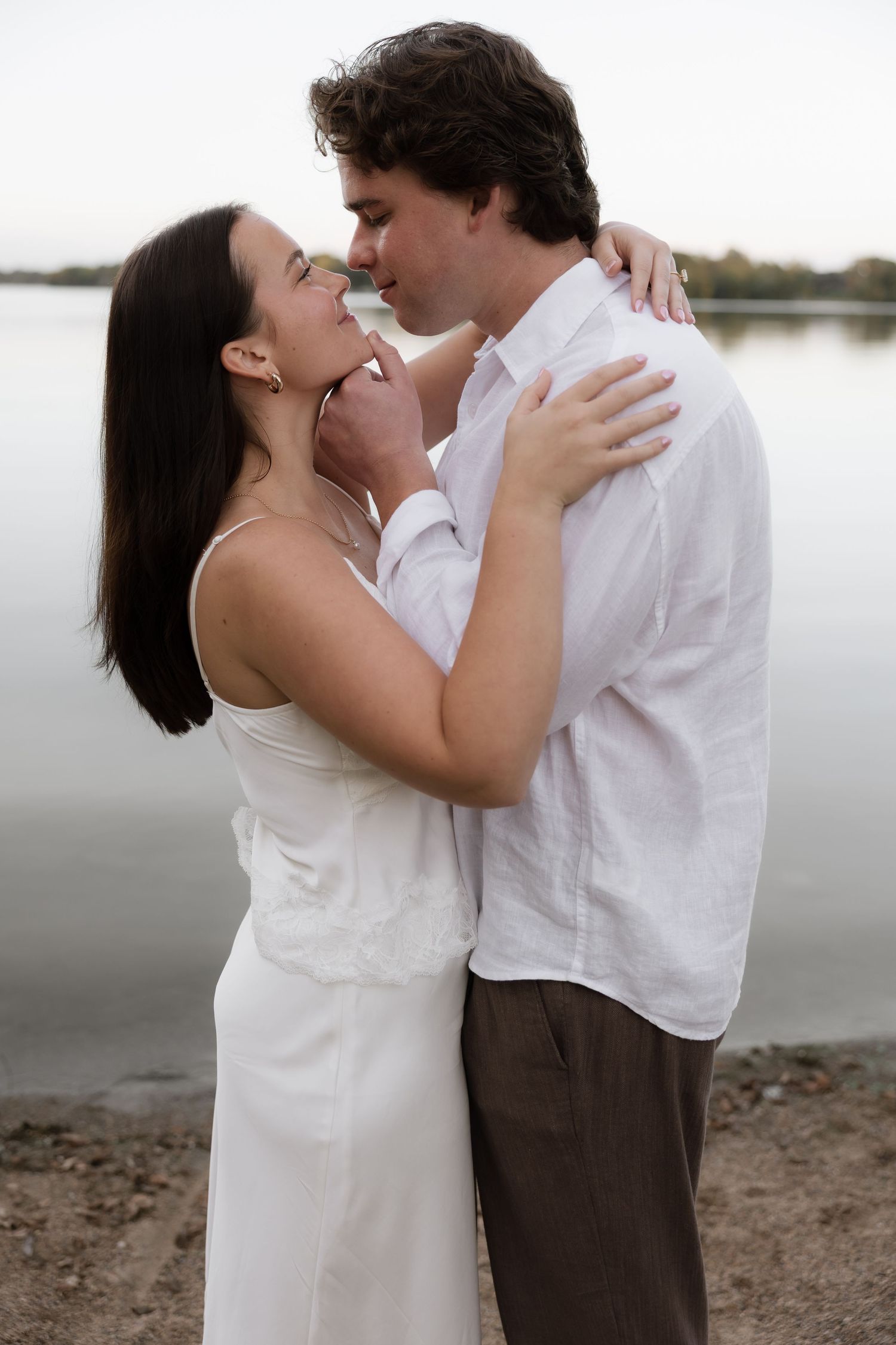Romantic couple embracing and almost kissing near a serene lake, woman in white dress, man in white shirt.