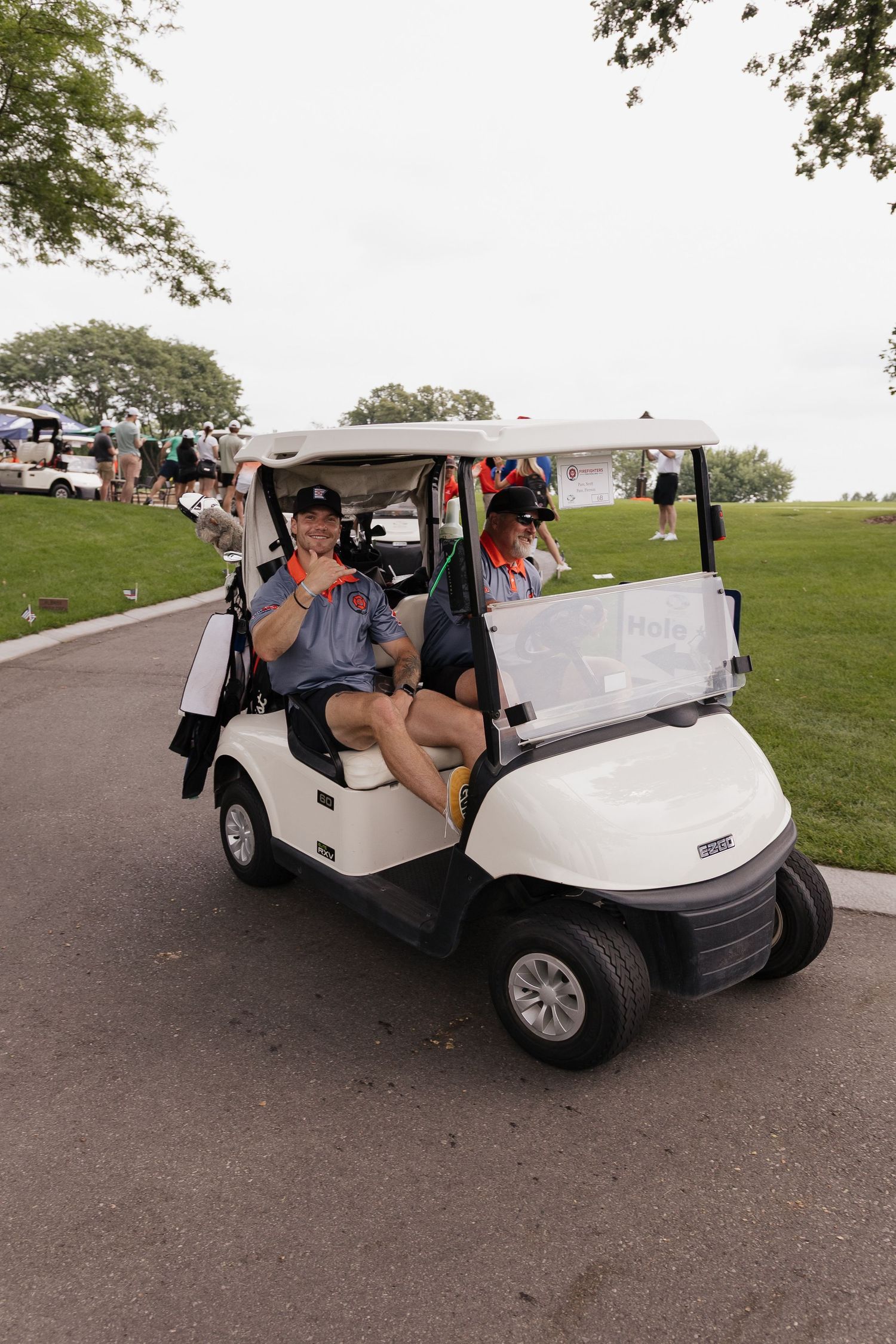 Two smiling people riding a white golf cart on a paved path at a golf course on a cloudy day.