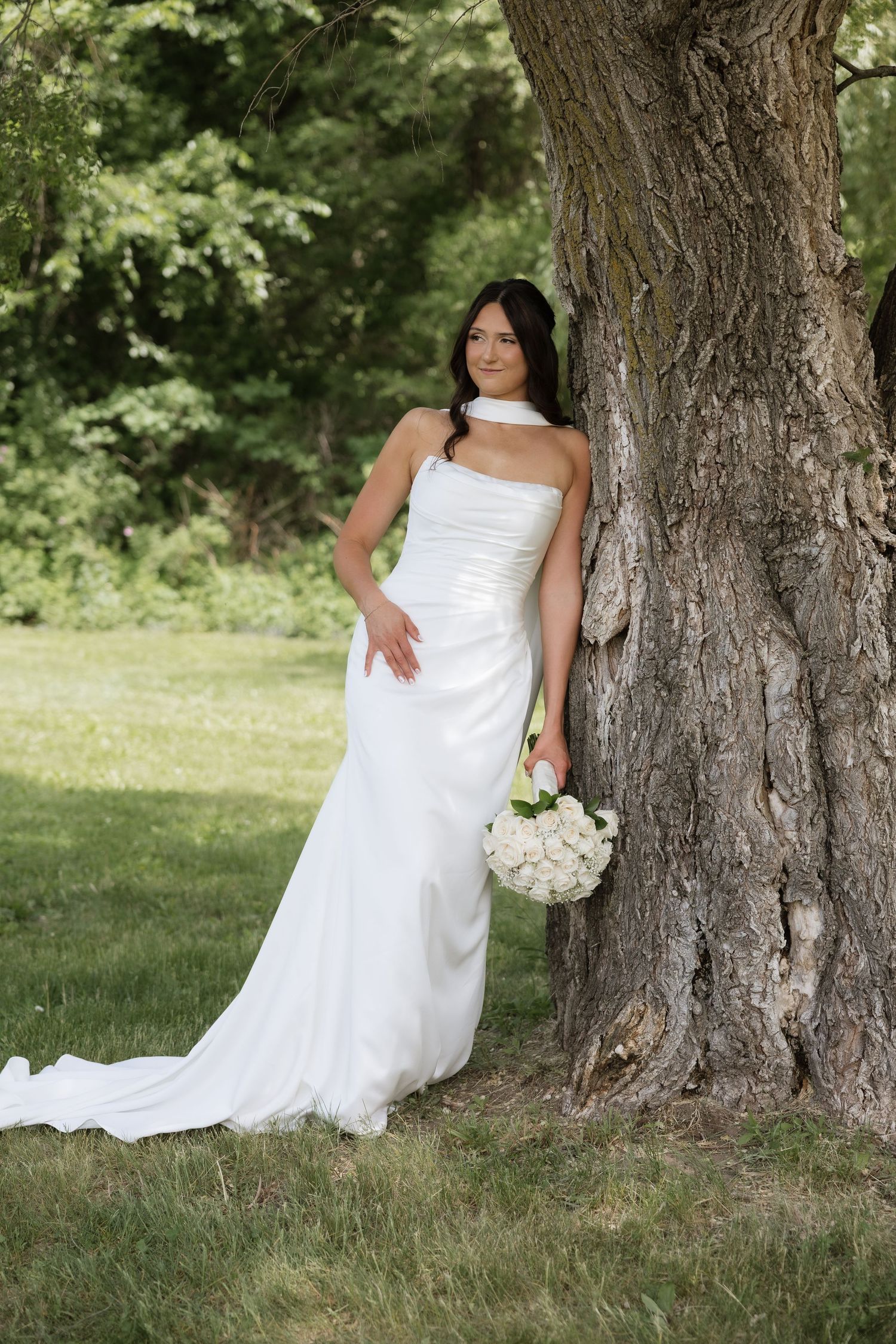 Elegant bride in a white halter wedding gown leans against a large tree while holding a white rose bouquet.