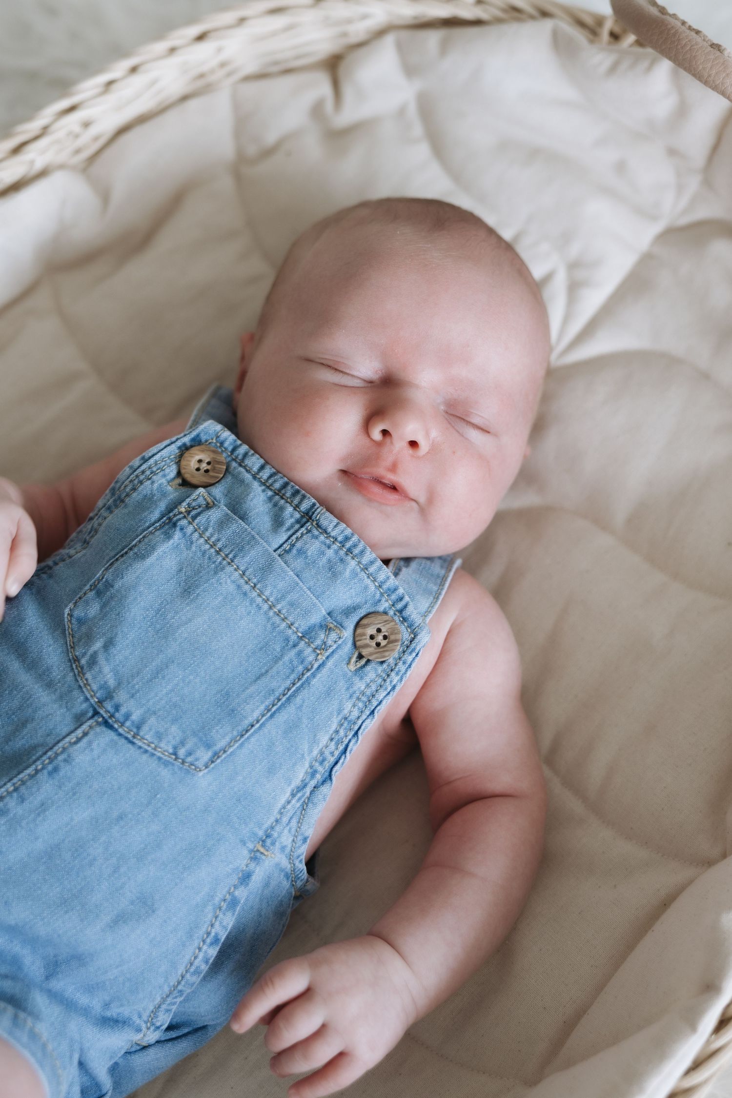 Peaceful newborn baby sleeping in wicker Moses basket wearing blue denim overalls.