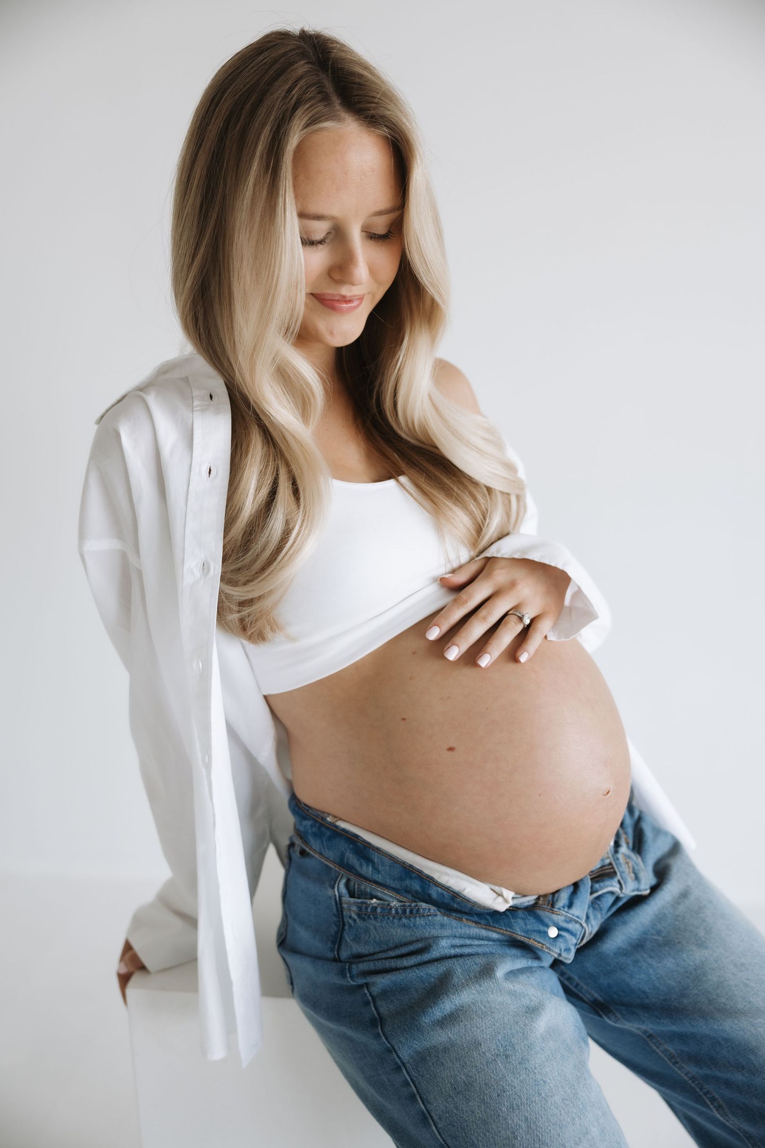 Smiling pregnant blonde woman in white crop top and jeans gently touching her baby bump.