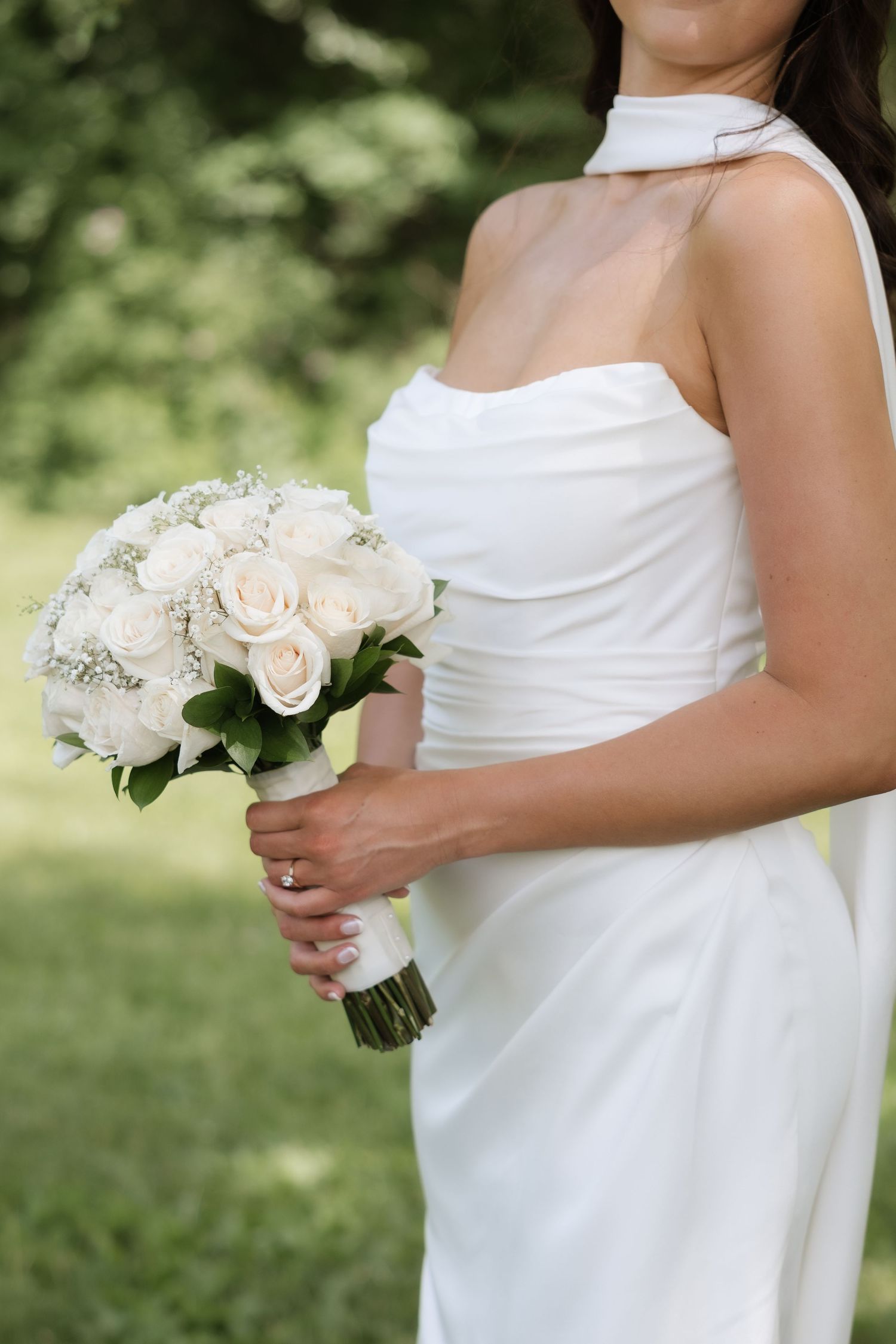 Bride in strapless white gown holding elegant white rose and baby's breath bouquet outdoors.