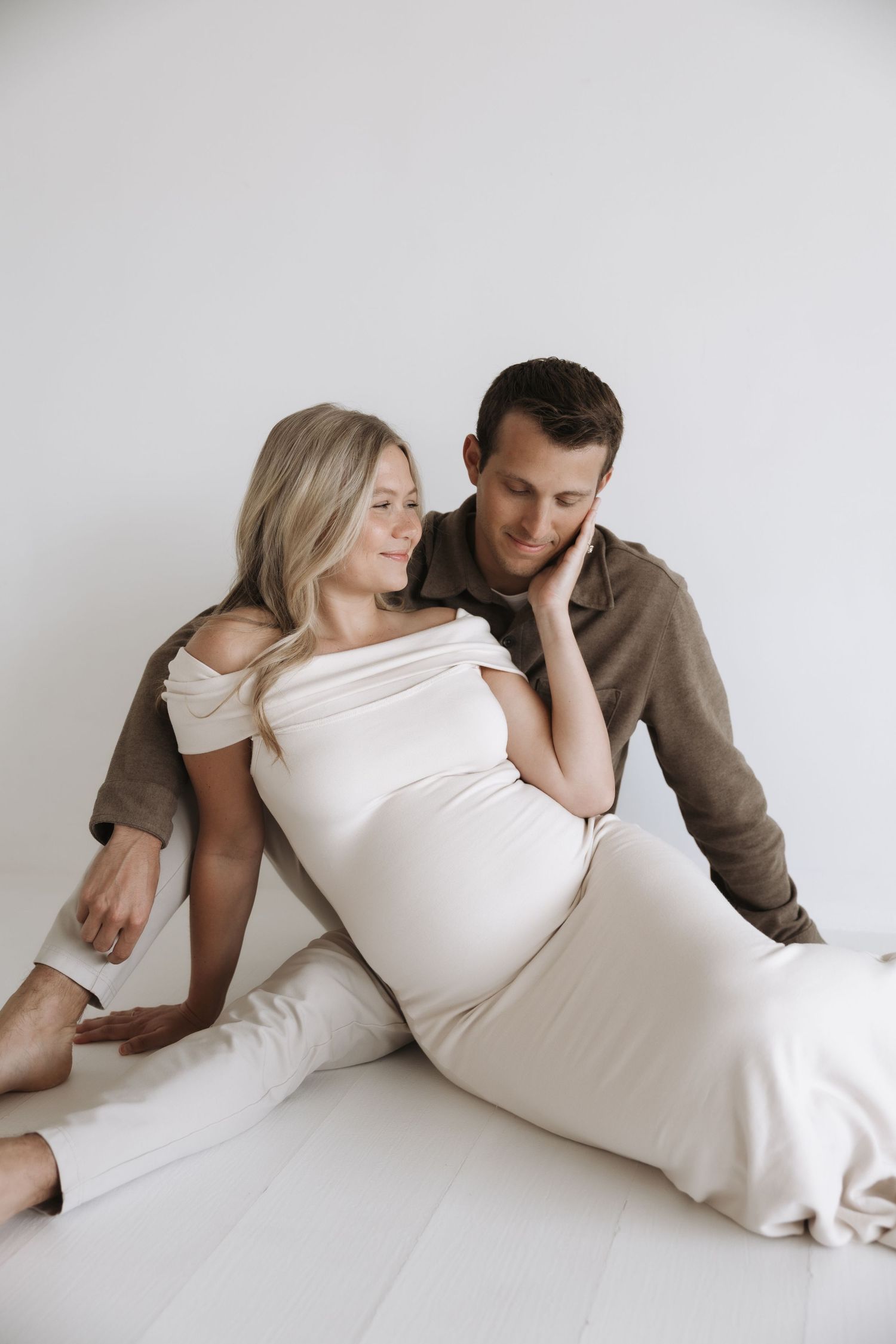 Couple sitting on floor during maternity shoot, woman in white dress leaning against partner.